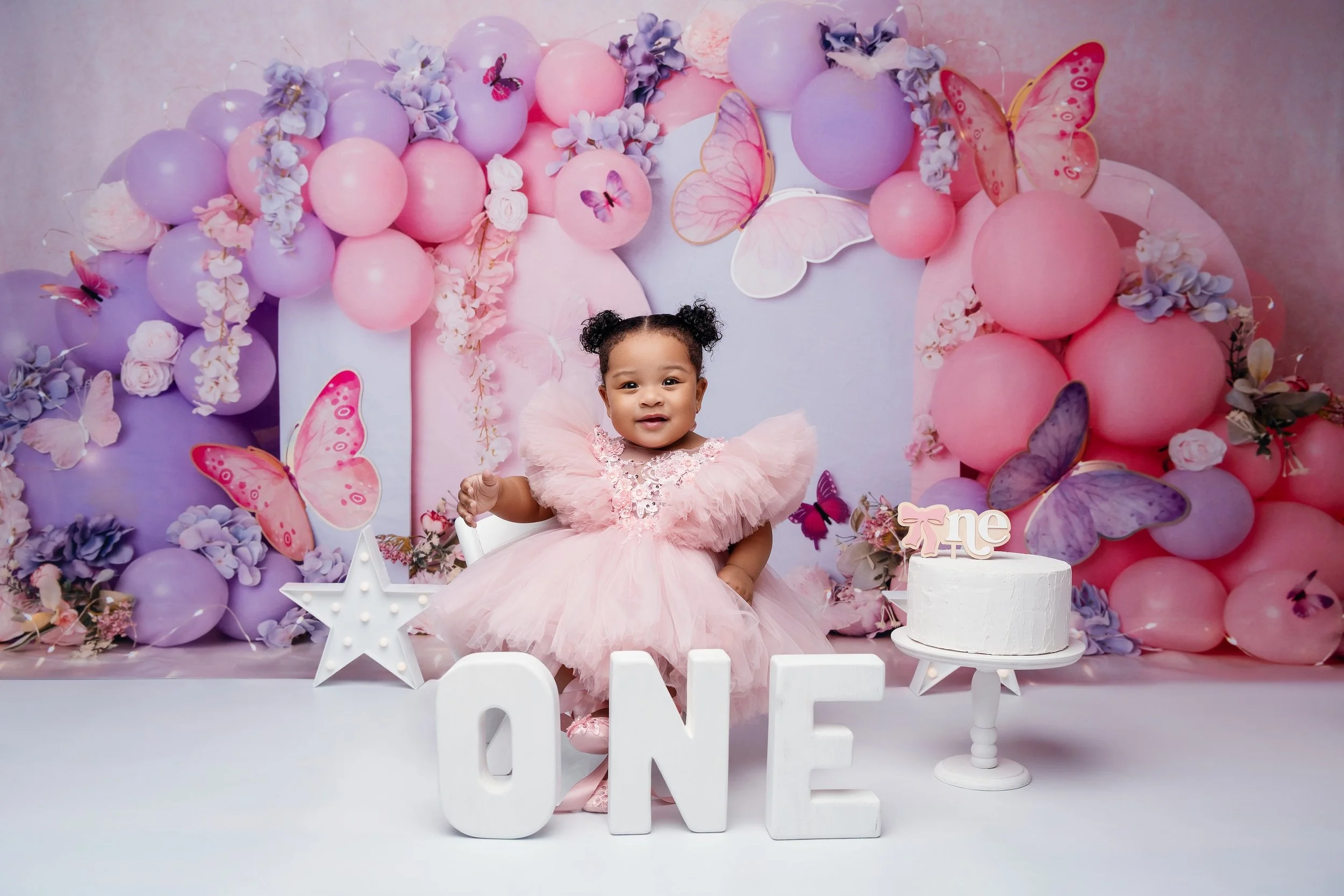 Baby girl in pink dress with butterfly balloon arch and birthday cake — vibrant cake smash studio setup for first birthday