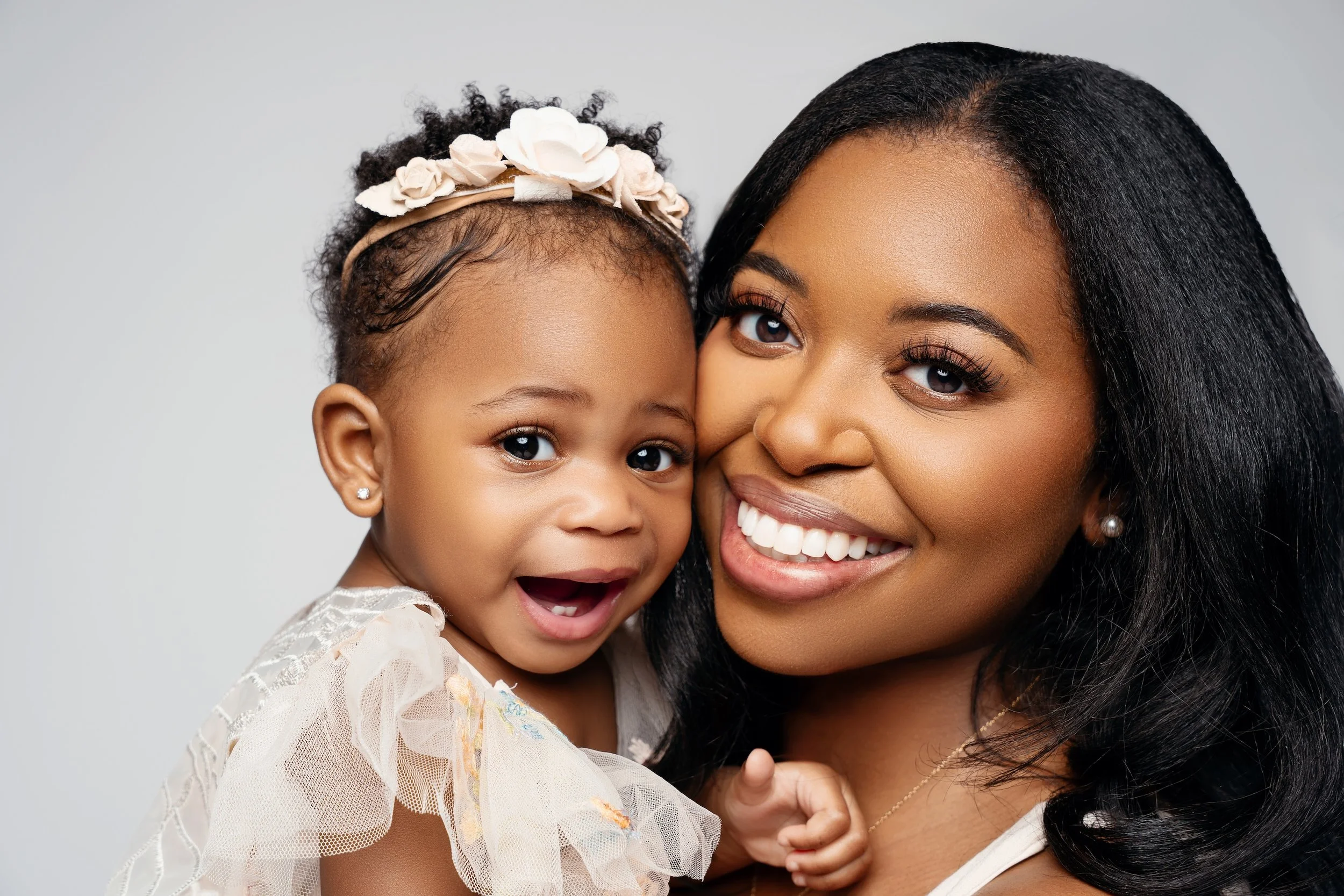Close-up portrait of a smiling mother holding her happy baby girl wearing a flower headband, captured by a luxury family photographer in a Chicago studio.