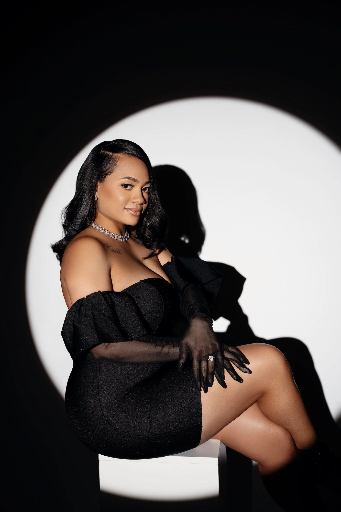 Solo spotlight portrait of engaged woman in black off-shoulder dress with sheer gloves and diamond jewelry, circle of light on black backdrop, Chicago studio