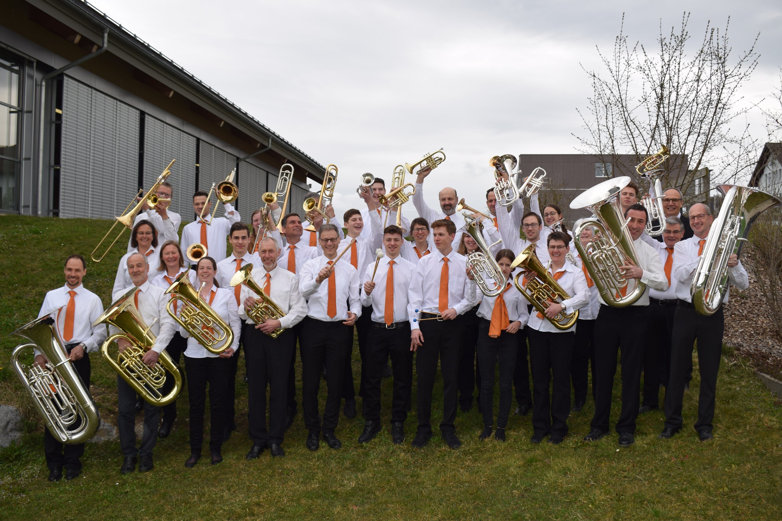 Eine Gruppe von Musikern mit Blasinstrumenten vor einem Gebäude und Bäumen im Hintergrund, alle tragen weiße Hemden und orange Krawatten.