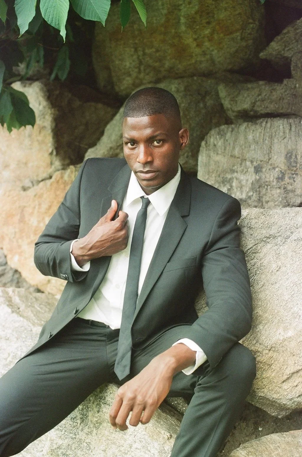 A man in a black suit and tie sitting on rocks outdoors, looking at the camera with his hand on his chest. There are large rocks and some green leaves in the background.