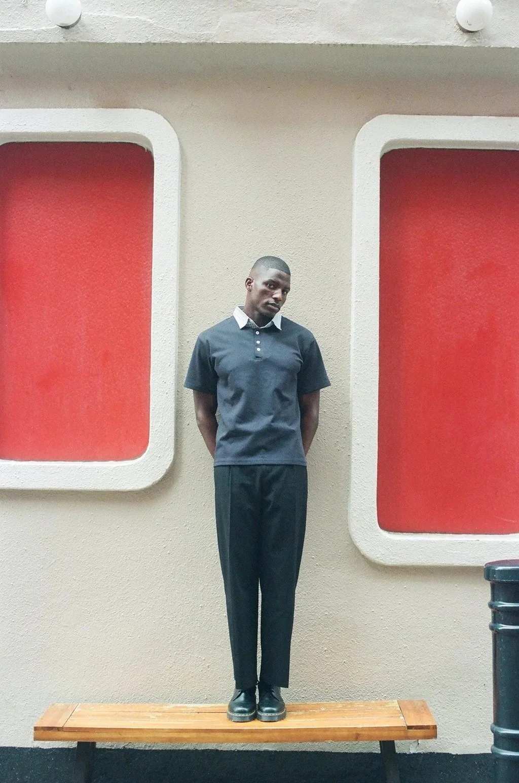 A young man in a dark polo shirt and black pants standing on a wooden bench in front of a beige wall with two large red square windows with white borders.