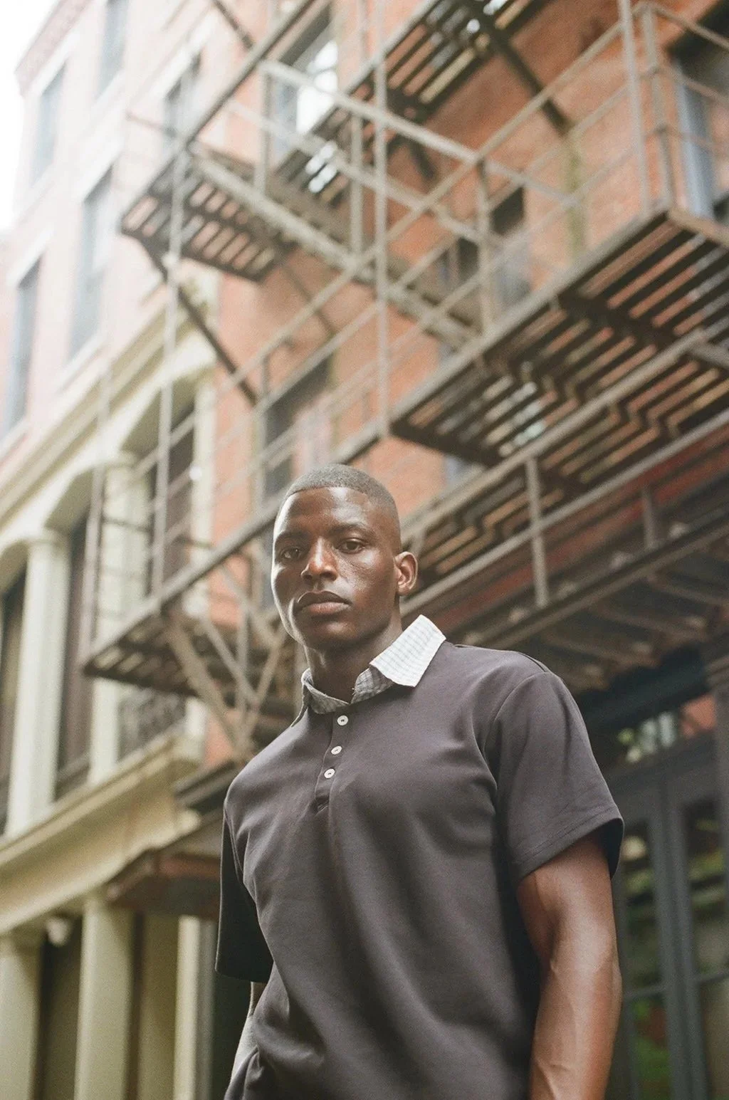 A young man standing outdoors in front of a brick apartment building with fire escapes, wearing a black polo shirt with a collared white shirt underneath.