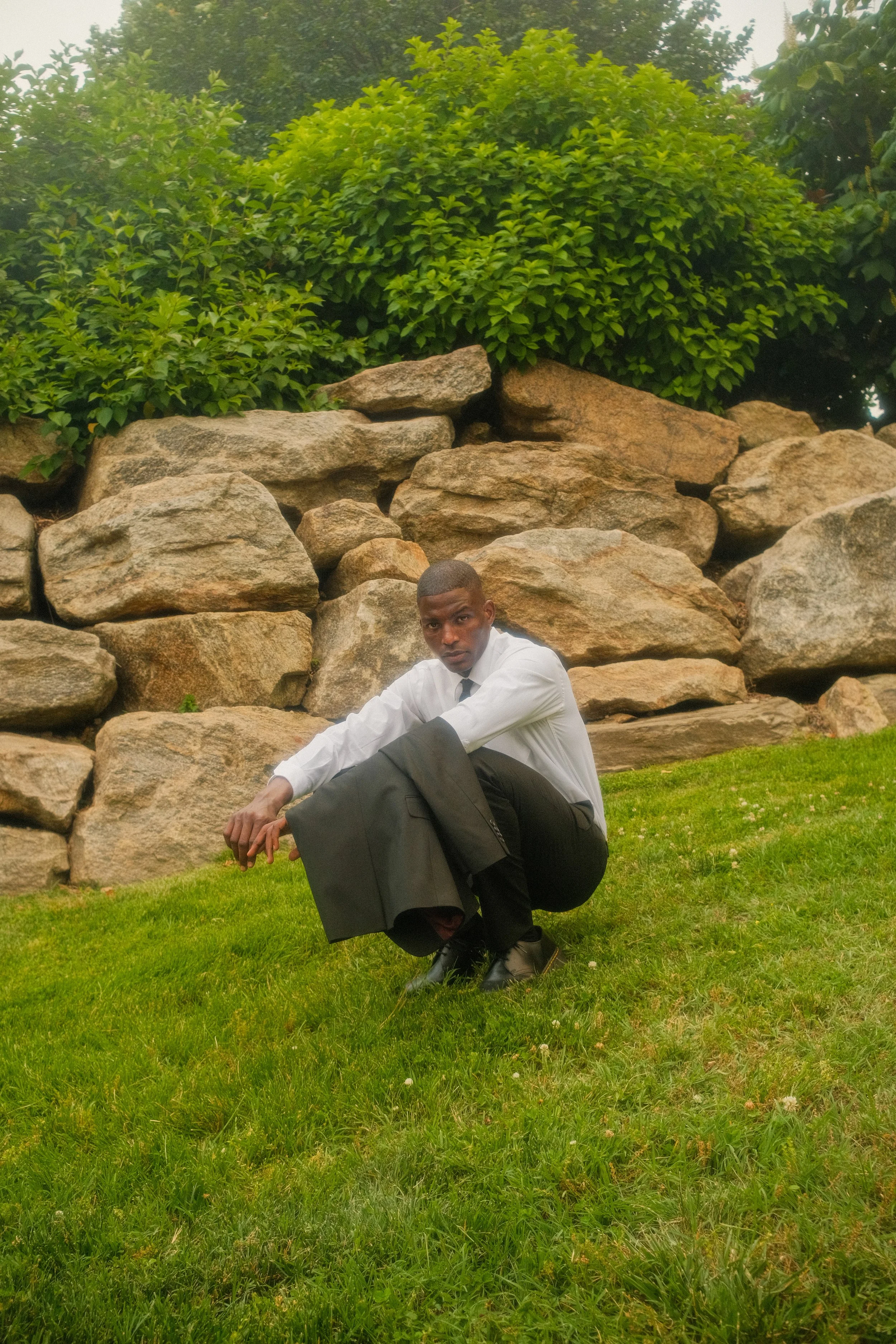 A man dressed in a white shirt and black tie, squatting on the grass in front of a rock formation with lush green bushes and trees in the background.