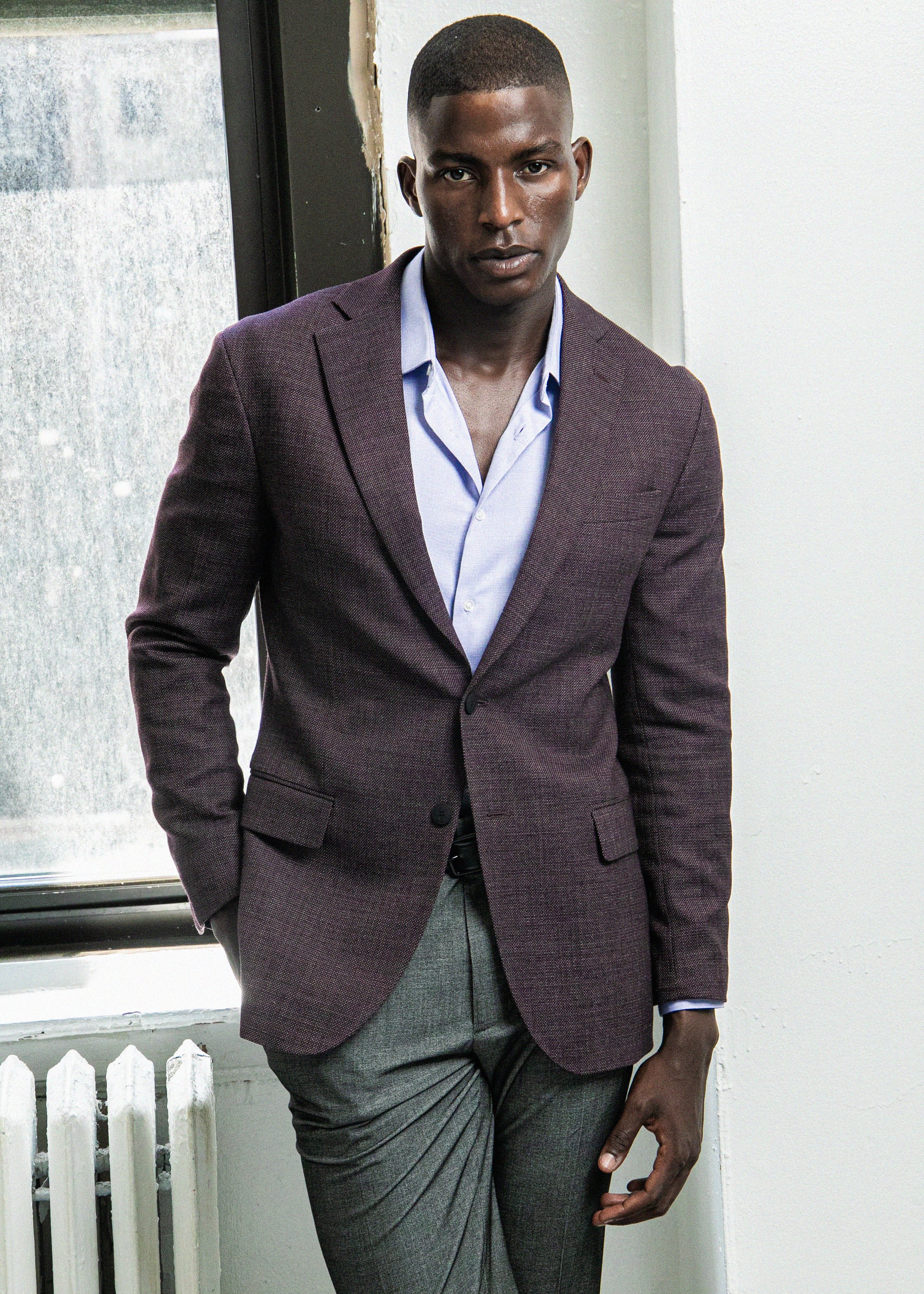 A man in a maroon blazer and grey trousers is standing indoors near a window with a white wall behind him. His right hand is pointing to his thigh.