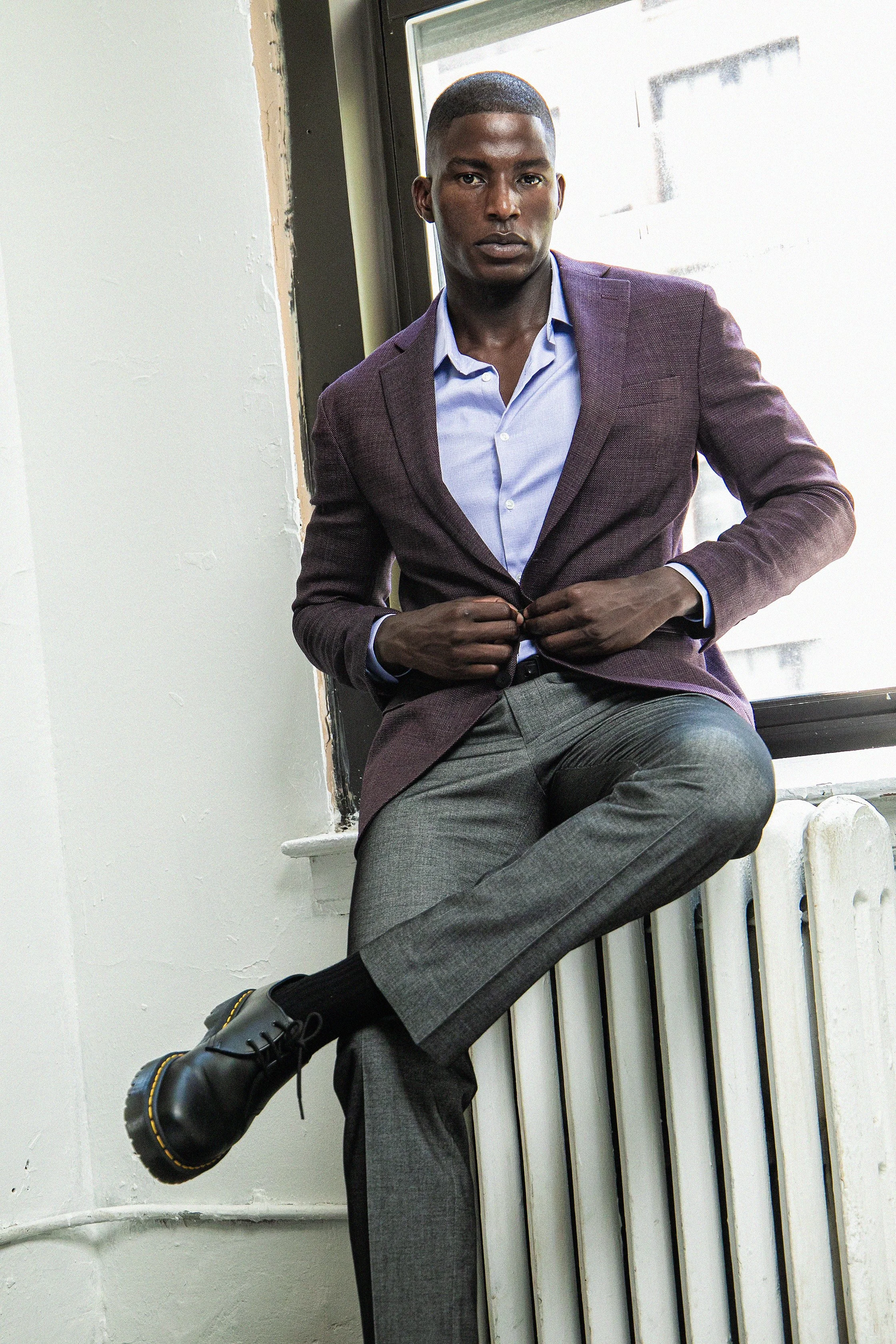 A young man with dark skin and short hair, wearing a purple blazer, light blue shirt, and gray pants, is sitting on a windowsill or radiator with one leg crossed over the other. He is looking directly at the camera with a serious expression.