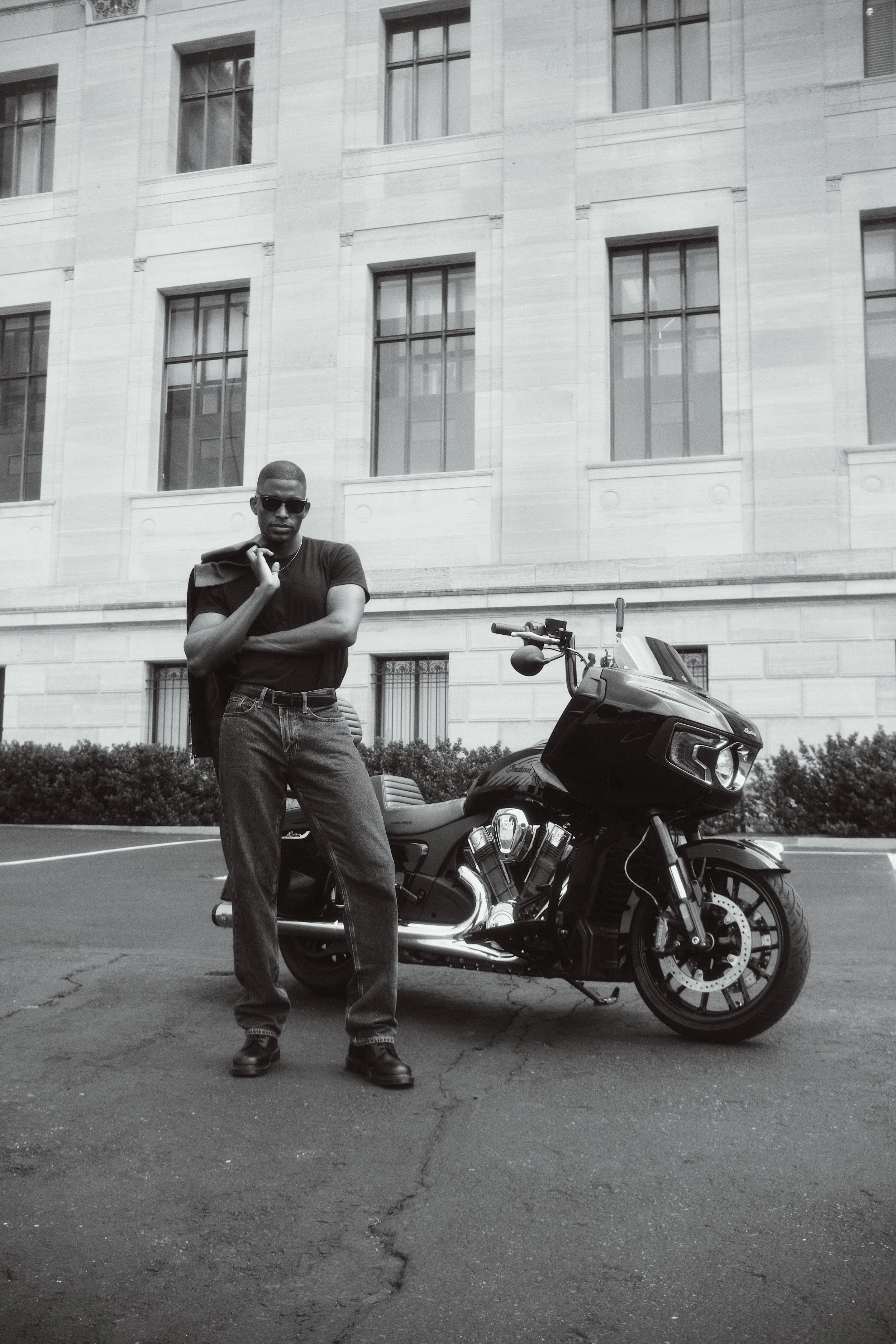 A man wearing sunglasses, a black t-shirt, and jeans standing next to a black motorcycle in front of a building with tall windows.