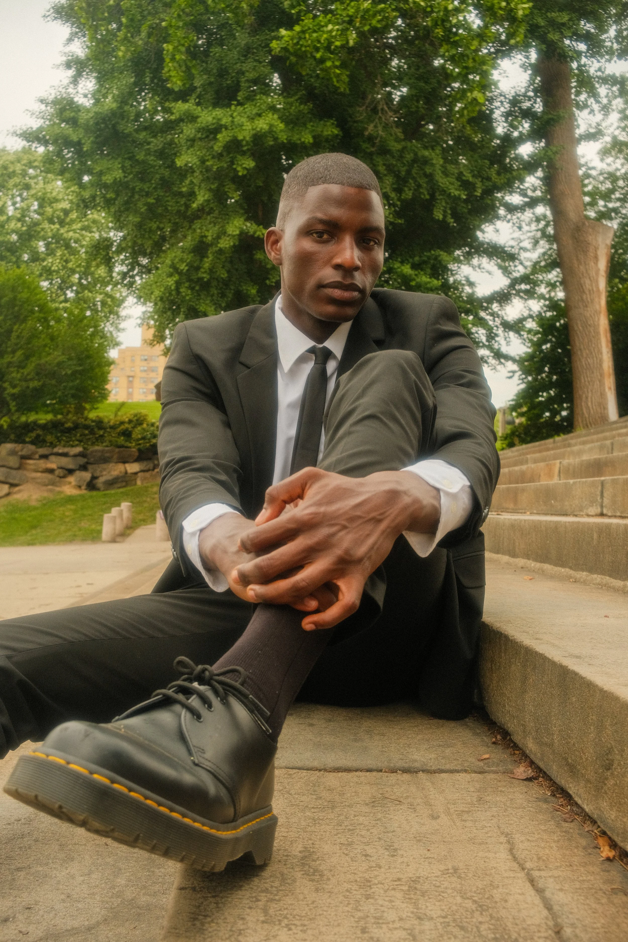 A young man dressed in a black suit, white shirt, and black tie sitting outdoors on concrete stairs with greenery and trees in the background. He has a serious expression and is wearing black shoes, with his left foot extended forward.