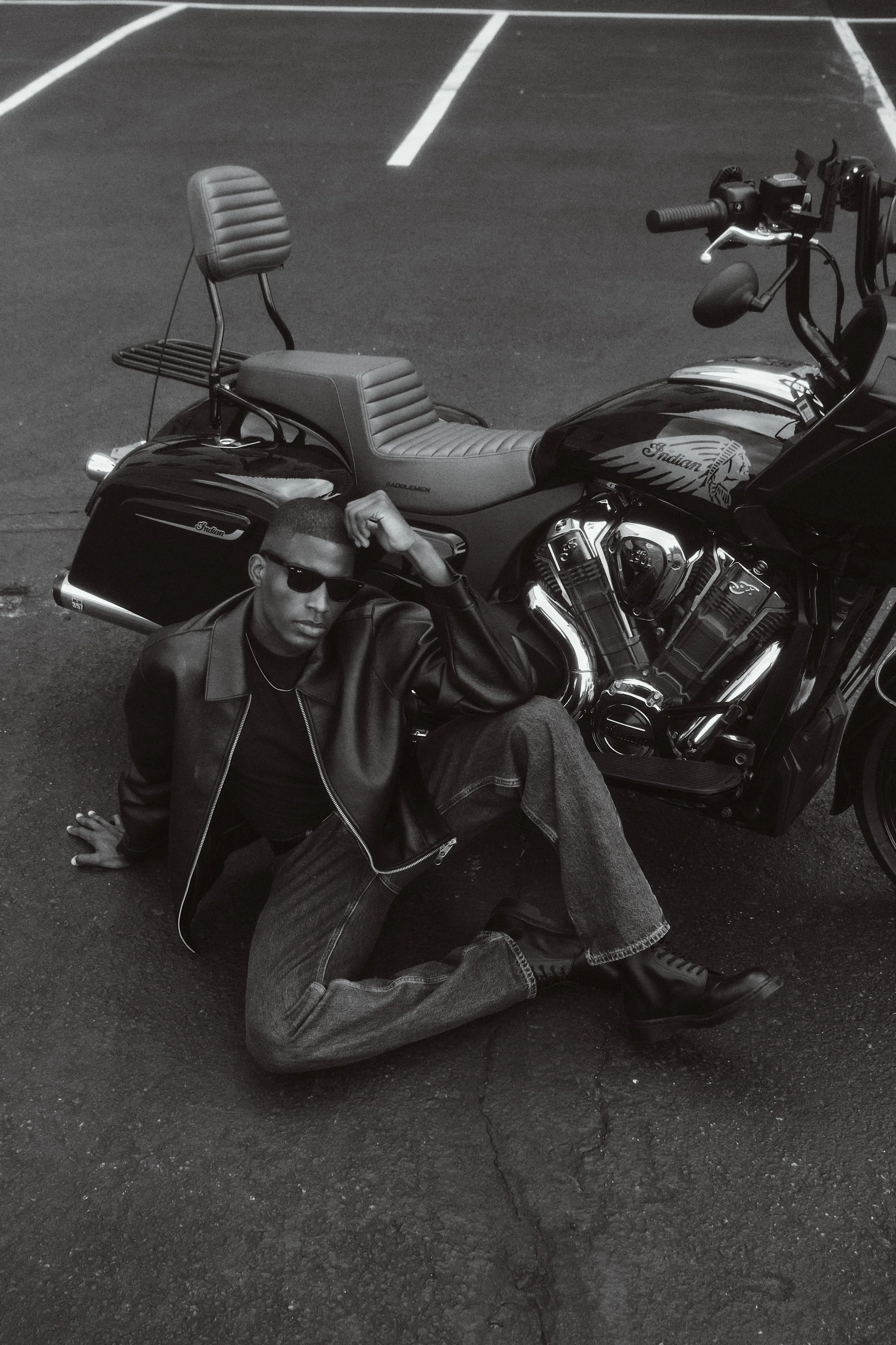 A man with sunglasses and a leather jacket is sitting on the ground next to a parked motorcycle with a large comfortable seat and detailed engine, in a parking lot with white parking space lines visible.