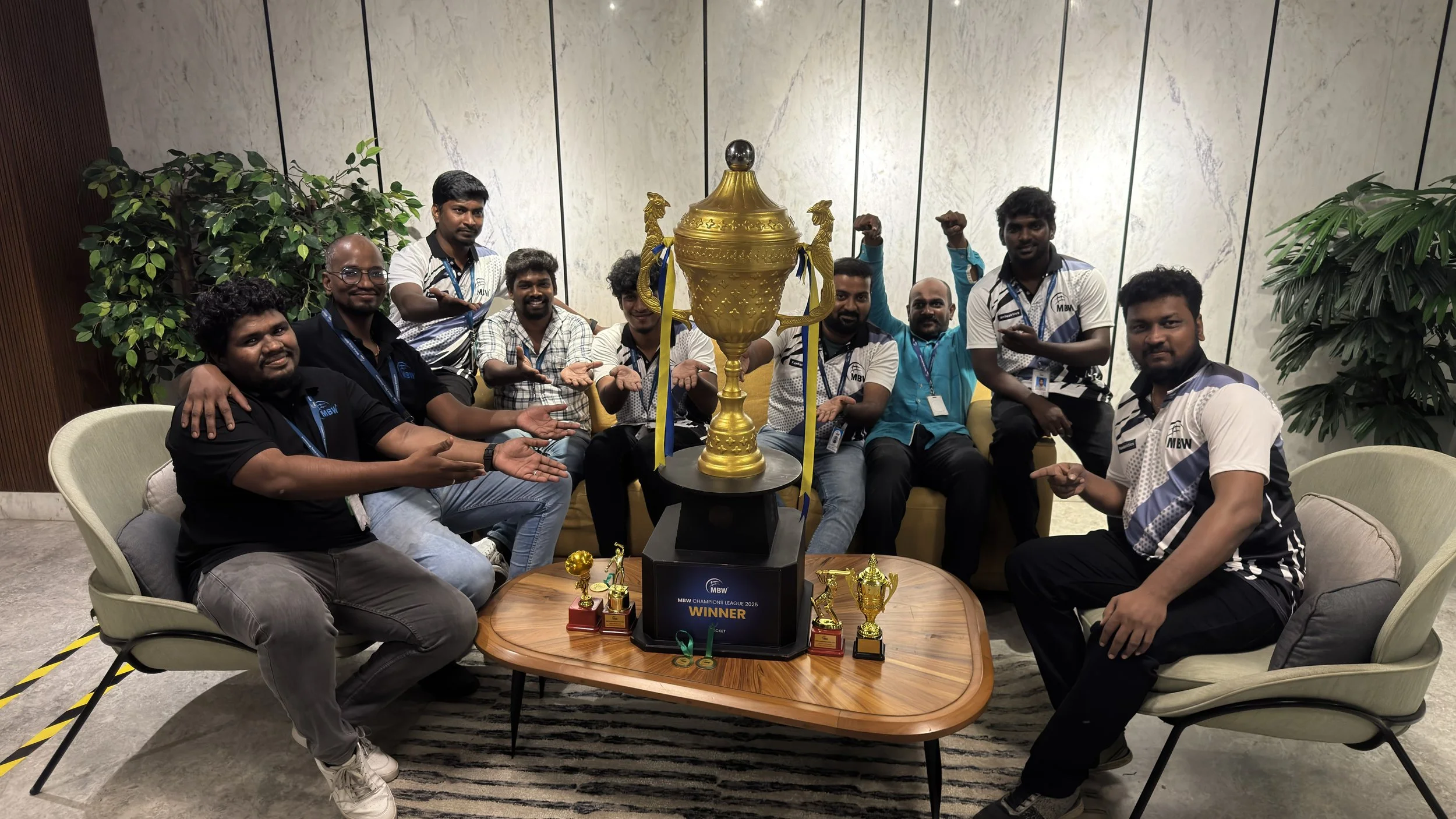 Group of nine men celebrating victory with a large trophy and smaller trophies on a wooden table, sitting and standing around in a modern indoor setting with plants and marble wall.