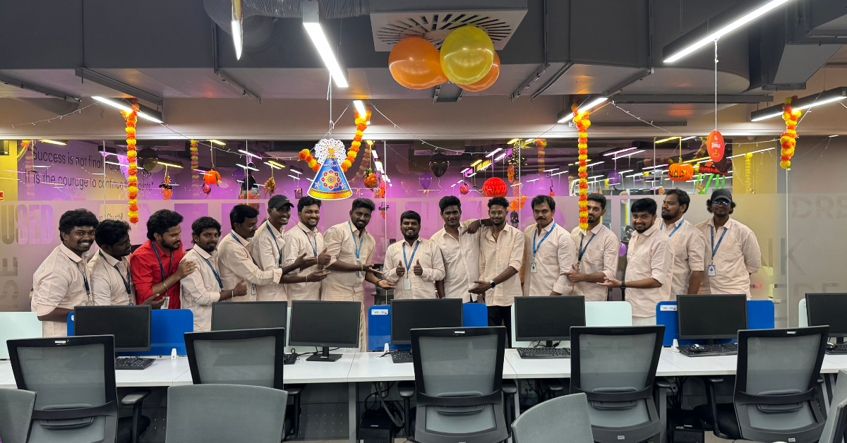 Group of colleagues standing together in an office decorated with balloons and hanging decorations for a celebration, with computer desks and chairs in the foreground.