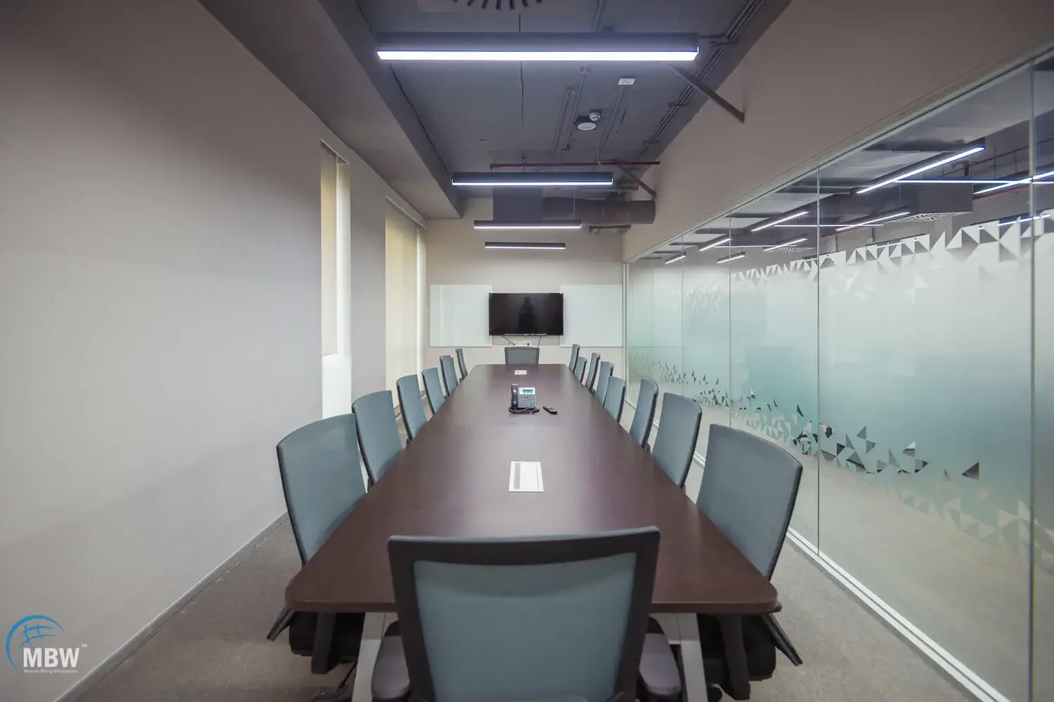 Empty conference room with a long table, chairs, a wall-mounted TV, a conference phone in the center, and windows with blinds.