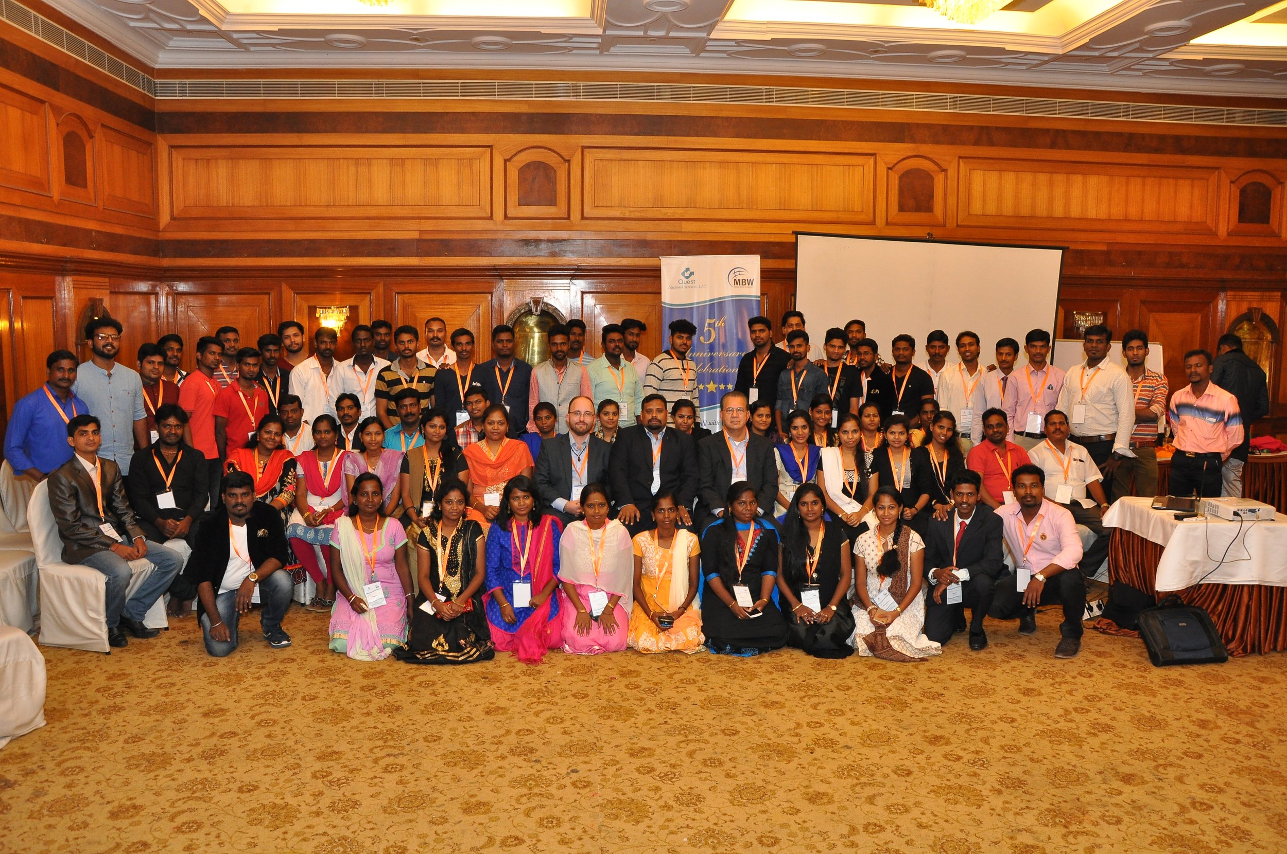 A large group of people gathered indoors for a conference or event, posing for a group photo in front of a wooden wall with a projector screen and a banner that reads '5th'.