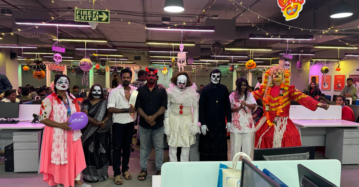 People dressed in Halloween costumes, including clowns, skeletons, and devils, posing in an office decorated with Halloween ornaments and lights.