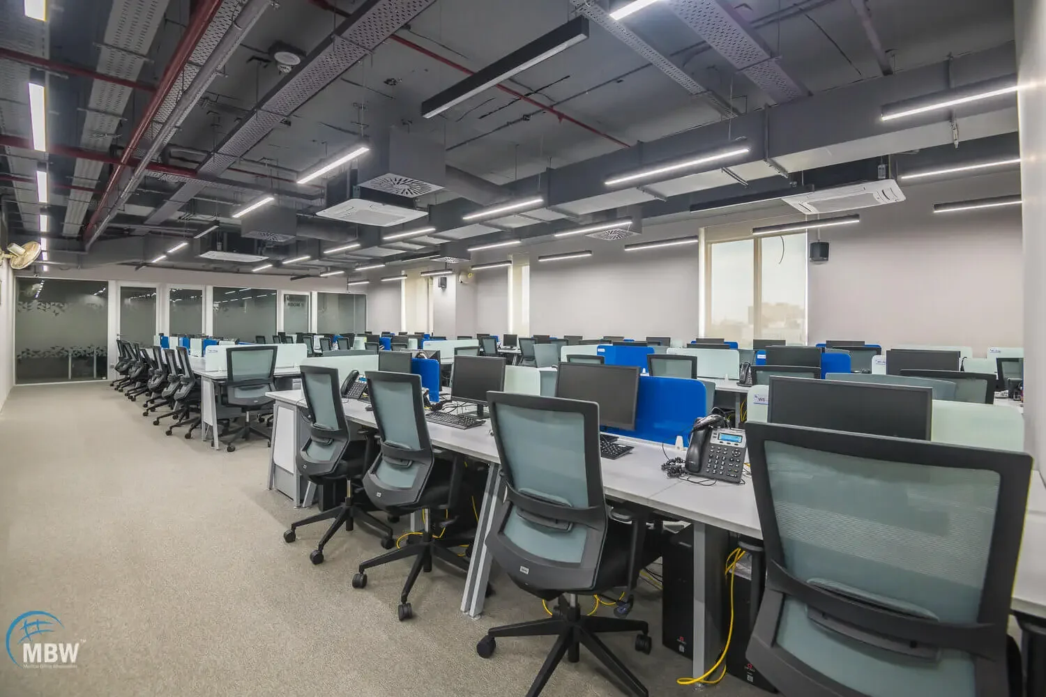 Modern office with multiple desks and computer workstations, black and gray chairs, and a ceiling with exposed ductwork and linear lighting fixtures.