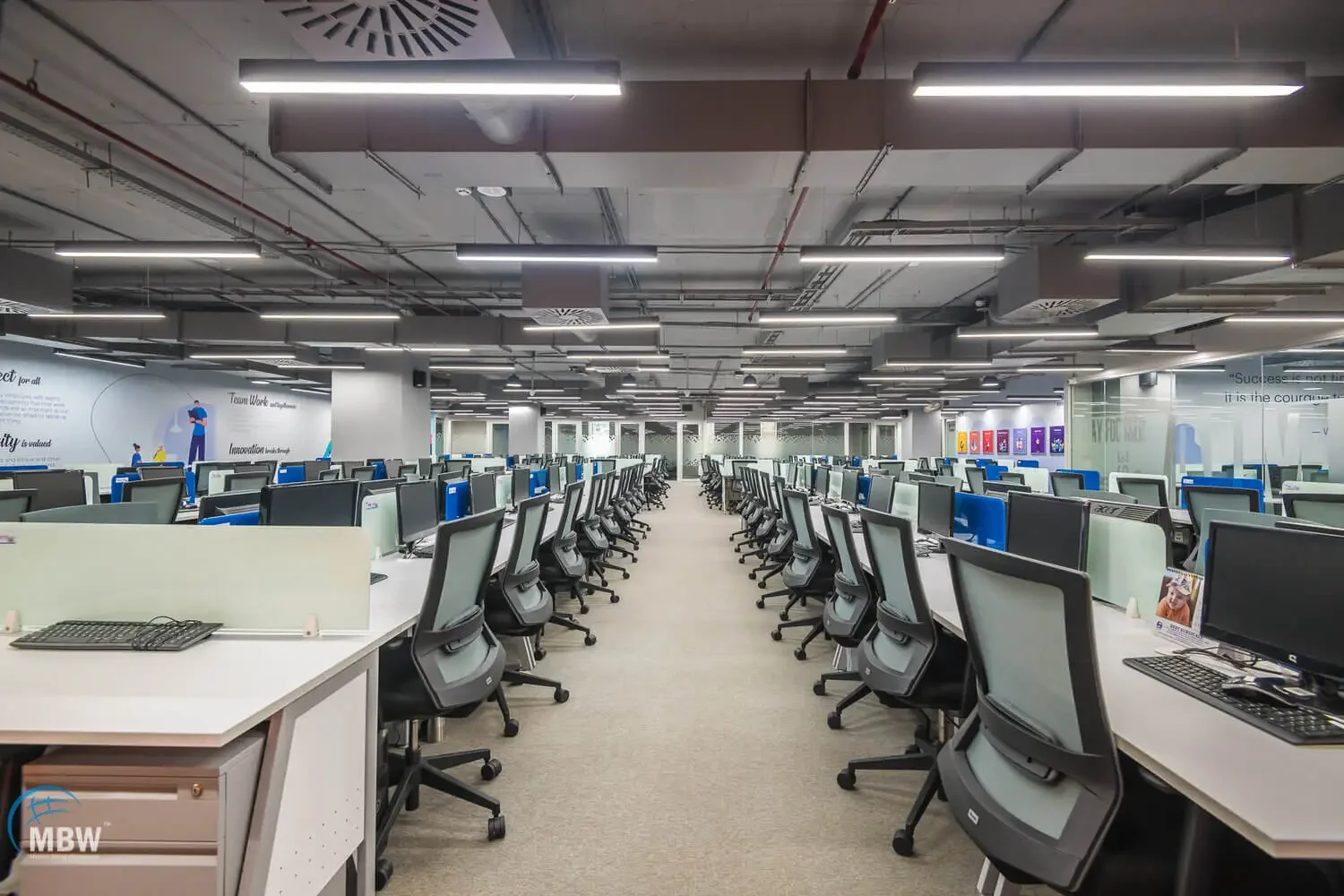 An empty modern office with rows of black office chairs and desktop computers, illuminated by overhead lighting, with glass walls and motivational signs on the walls.