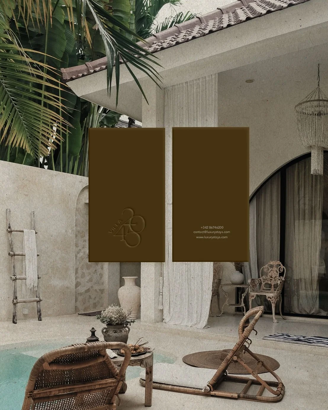 A cozy outdoor patio area with wicker chairs and a small table near a swimming pool, surrounded by lush green plants and elegant decor, with two brown vertical banners displaying the logo, contact information, and website of Vila 45 luxury stay.