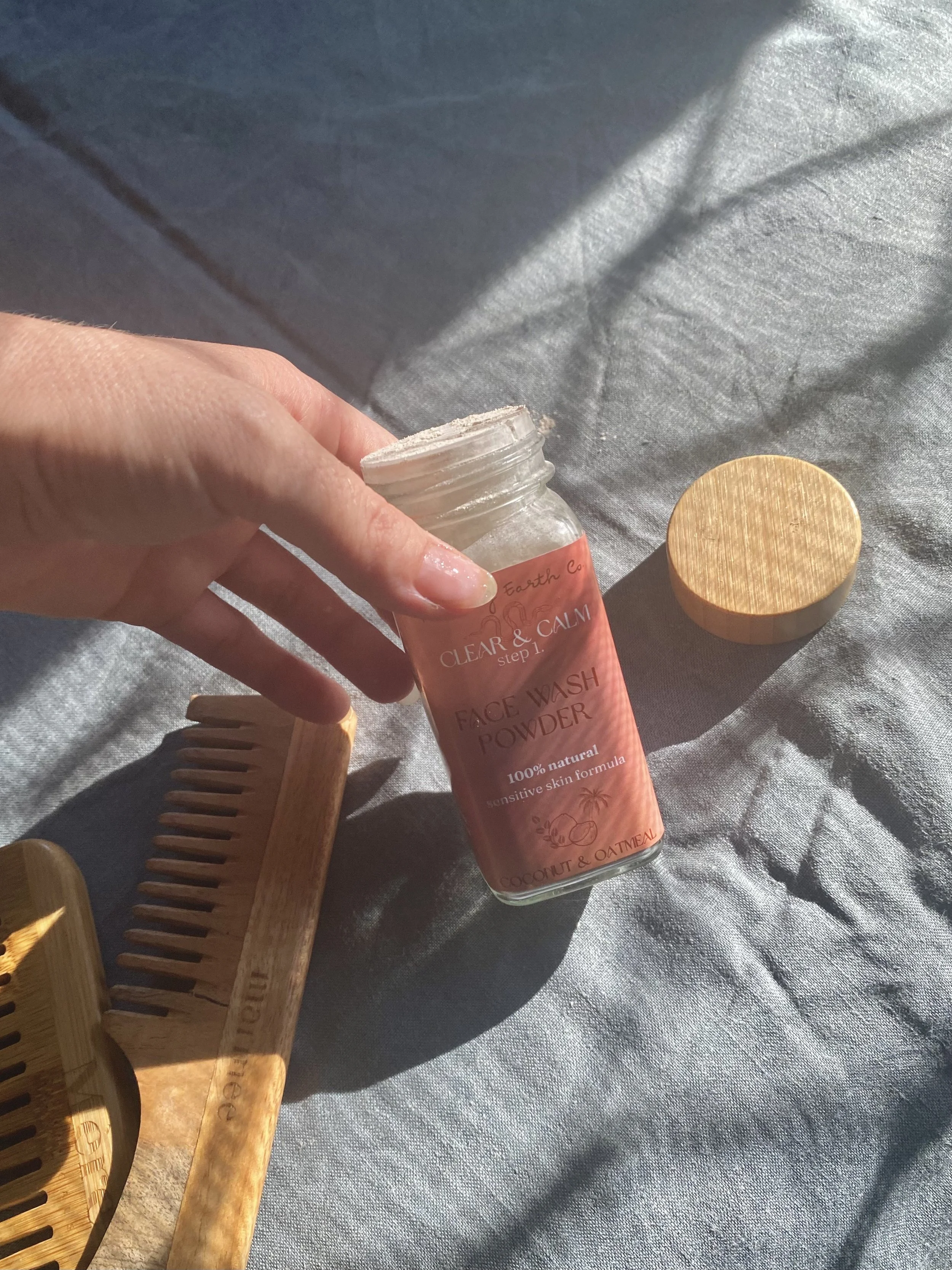 A person holding a jar of face wash powder labeled 'Earth Co. Clear & Calm Step 1 Face Wash Powder, 100% natural, sensitive skin formula, coconut & oatmeal.' The jar lid is placed on a gray fabric surface, alongside a wooden comb and a wooden lid.