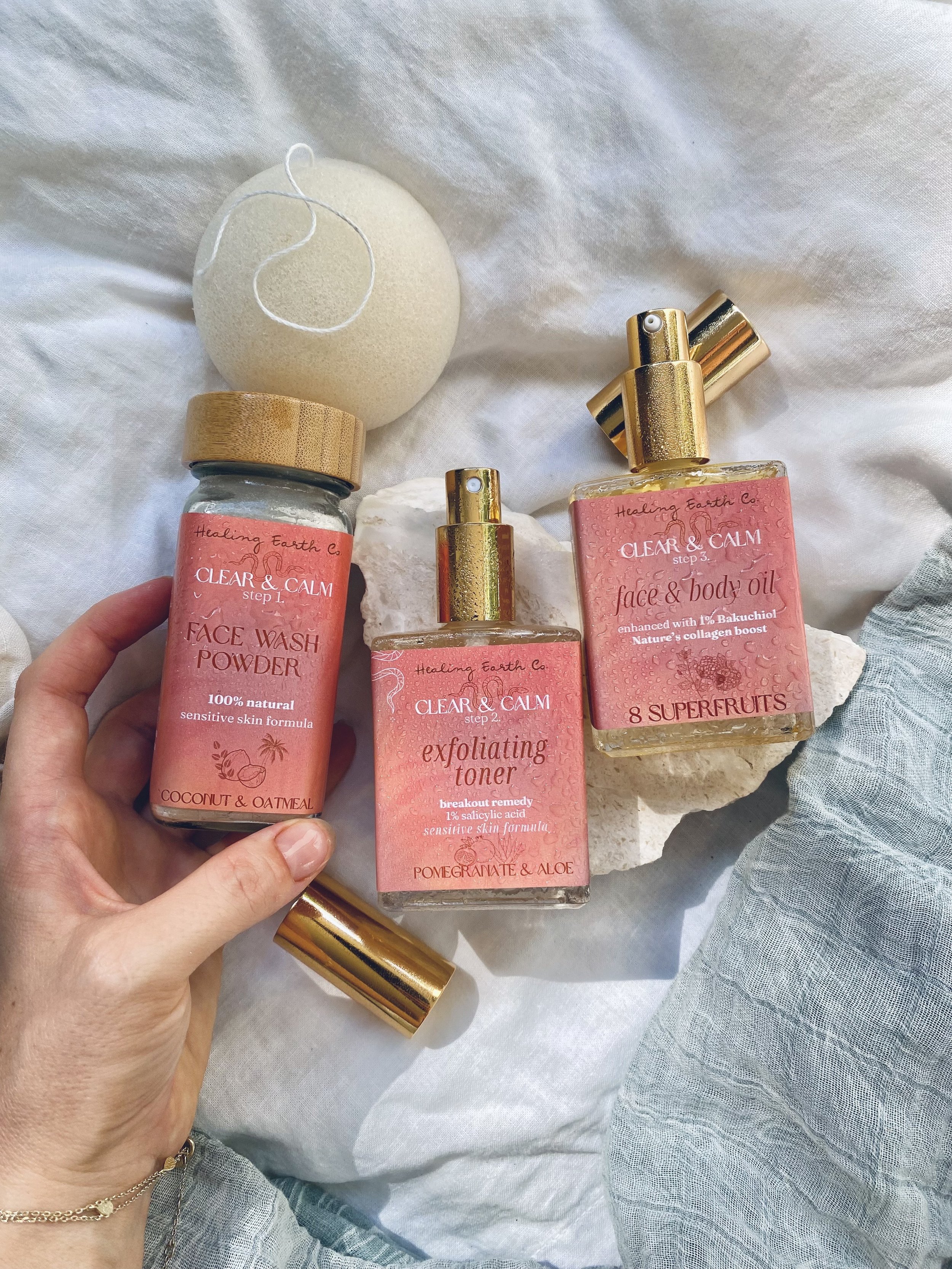 A hand holding a small container of face wash powder among skincare products, with a soap sponge and rocks on a soft, white fabric background.