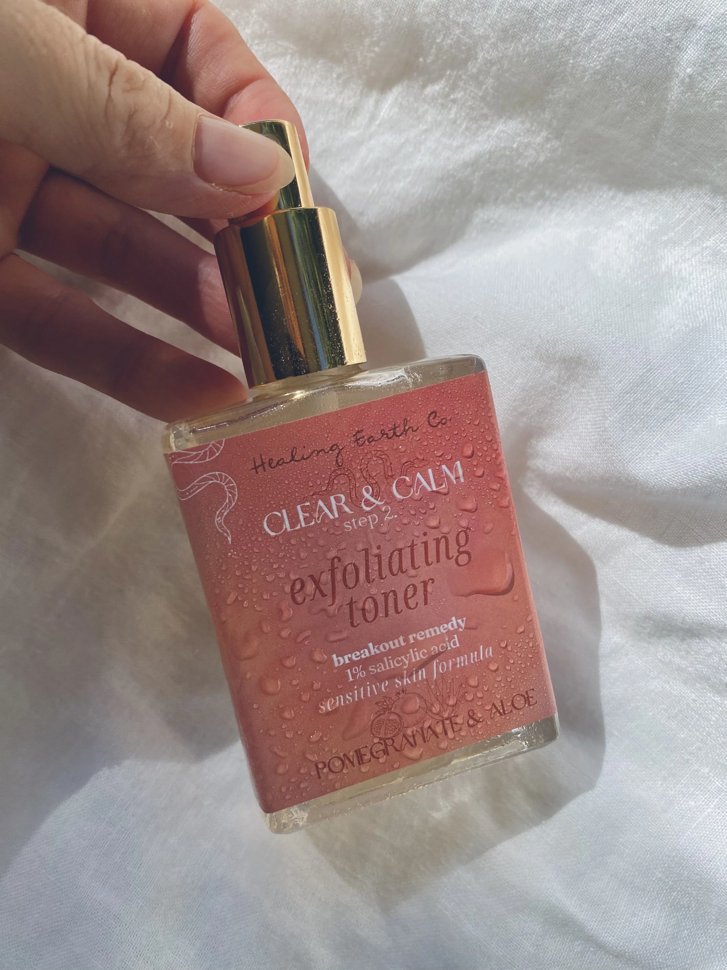 Hand holding a glass bottle of exfoliating toner with a pump dispenser, label reads 'Clear & Calm exfoliating toner, breakthrough remedy, 1% salicylic acid, sensitive skin formula, pomegranate & aloe, Healing Earth Co.' on a white fabric background.