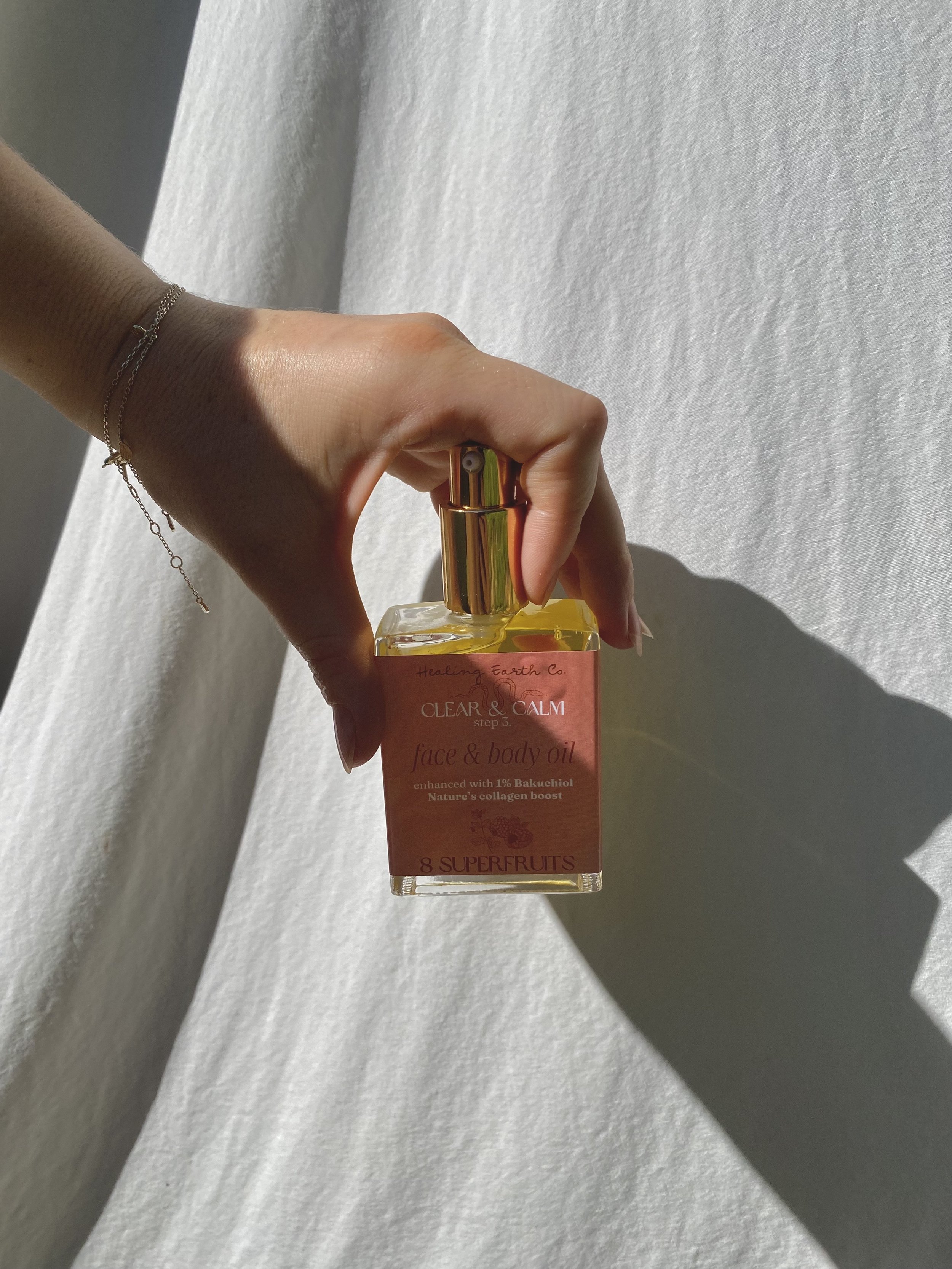 Hand holding a small glass bottle of face and body oil with a pink label, against a white background.