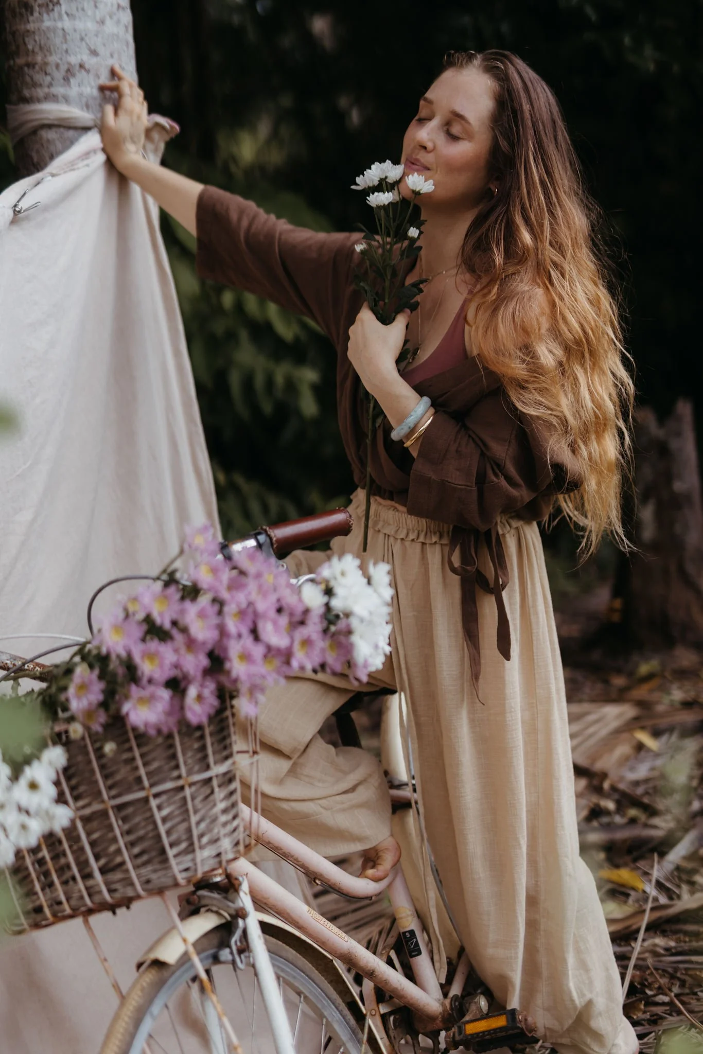 A woman with long, curly hair holding white daisies near her face while standing outdoors next to a bicycle with a basket of pink and white flowers.