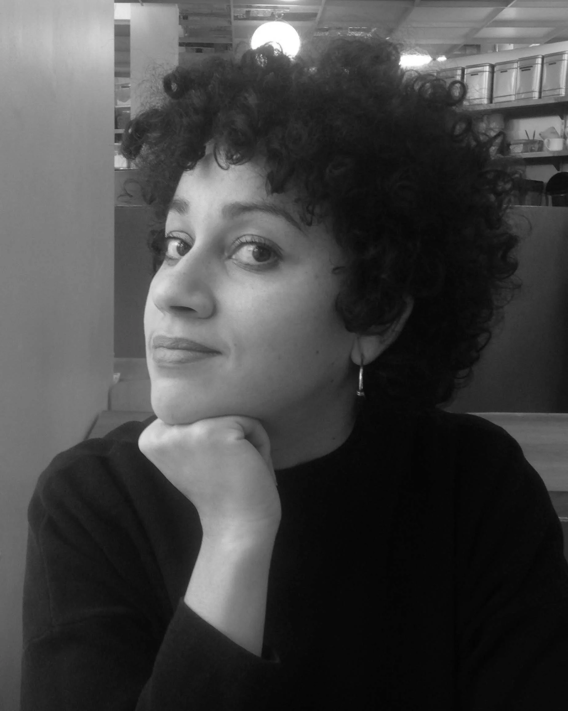 Black and white photo of a woman with curly hair, resting her chin on her hand, looking at the camera with a slight smile, in a indoor setting with shelves in the background.
