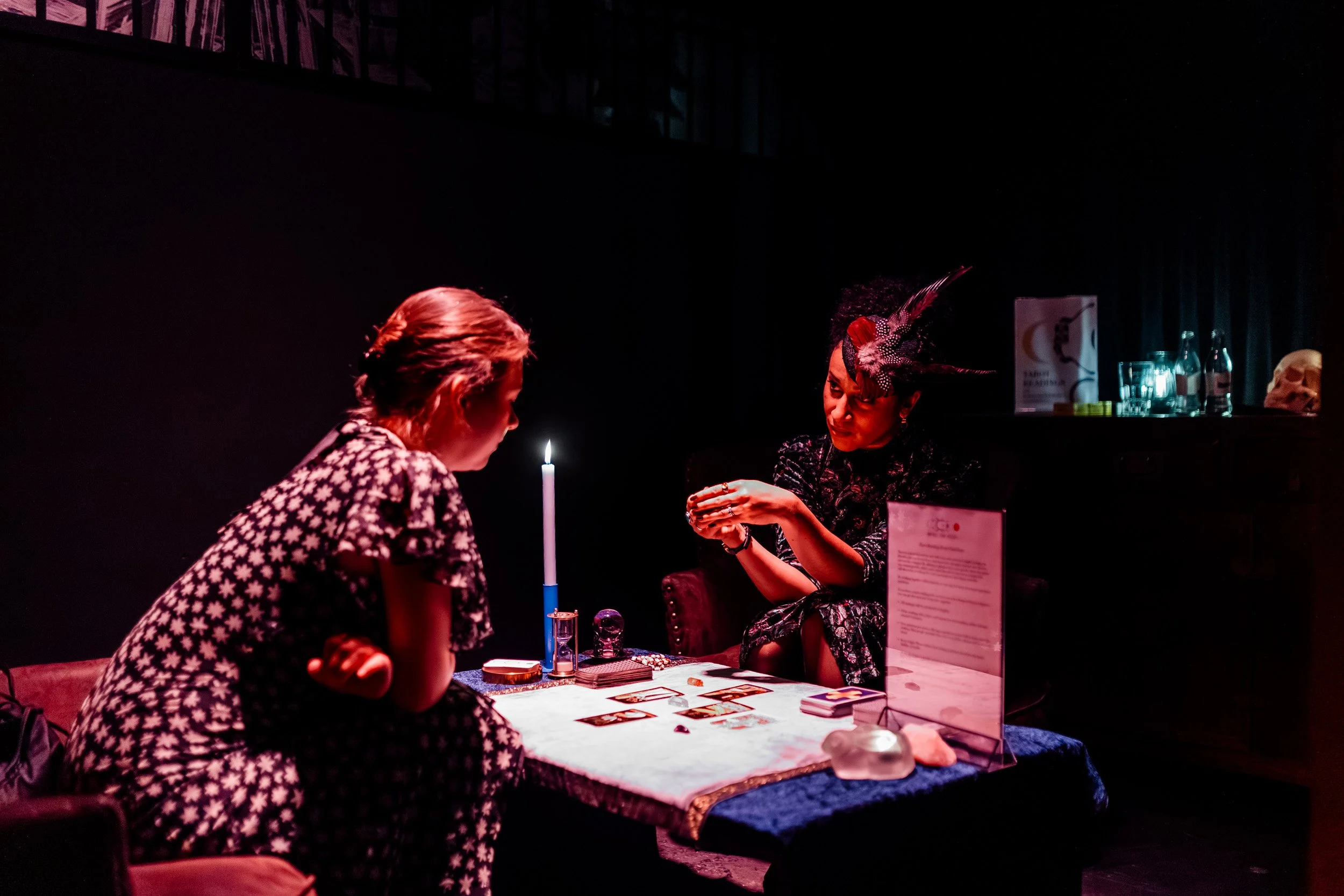 Two women sitting at a table in a dark room, playing tarot cards with a lit candle between them, one woman wearing a patterned dress and the other with an elaborate headpiece.