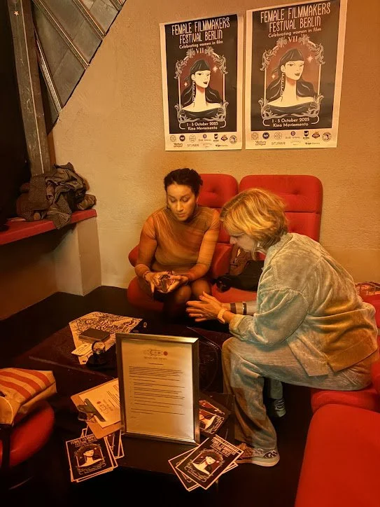 Two women sitting on red chairs in a room decorated with posters for the Female Filmmakers Festival Berlin, 2005. One woman with dark hair and an orange top is looking at her phone; the other with blonde hair and a gray jacket is showing her something. A small table in front of them has various brochures, flyers, and a framed document.