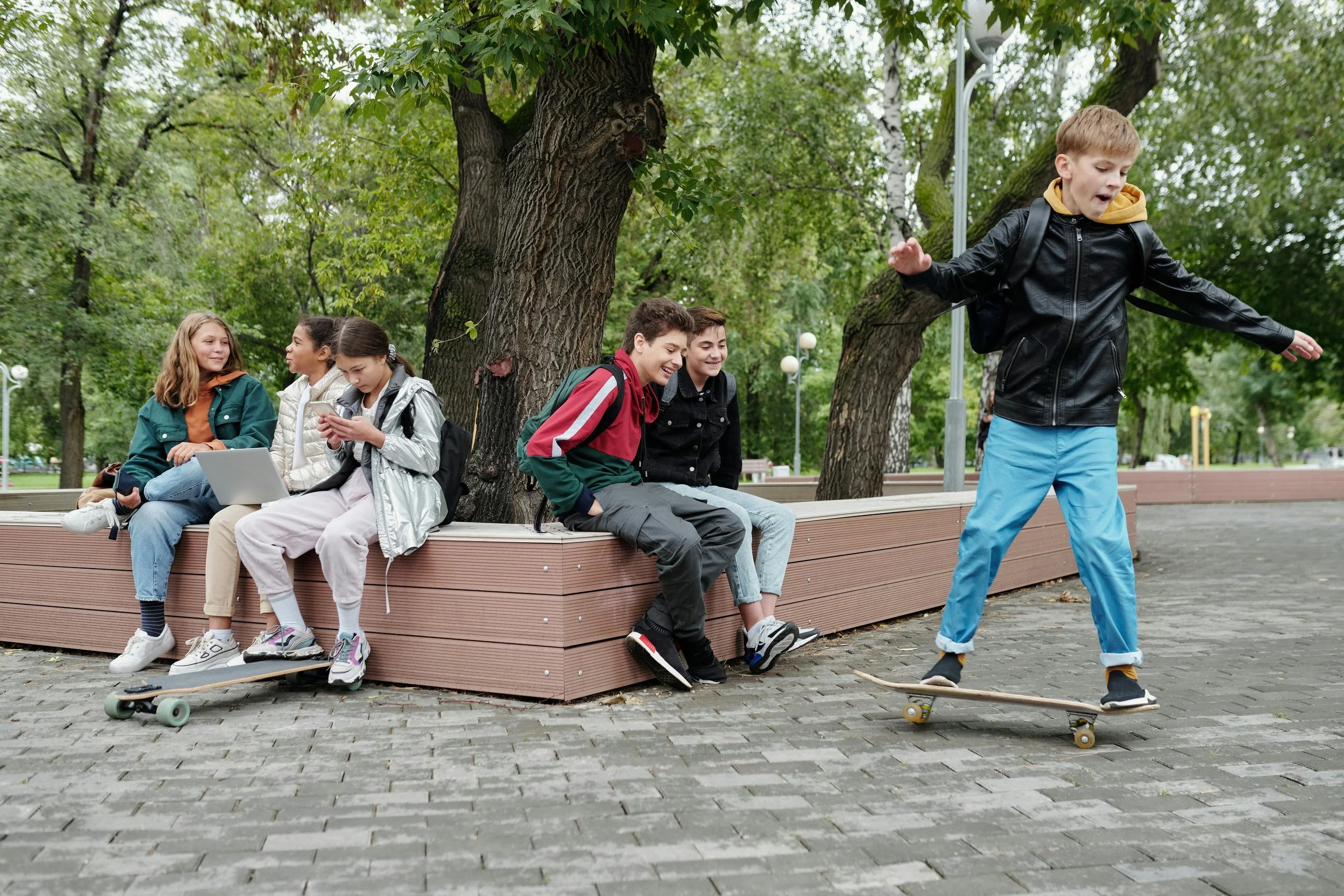 Four girls sitting on a park bench with backpacks, one girl on a skateboard, and a boy skateboarding on cobblestone path surrounded by trees.