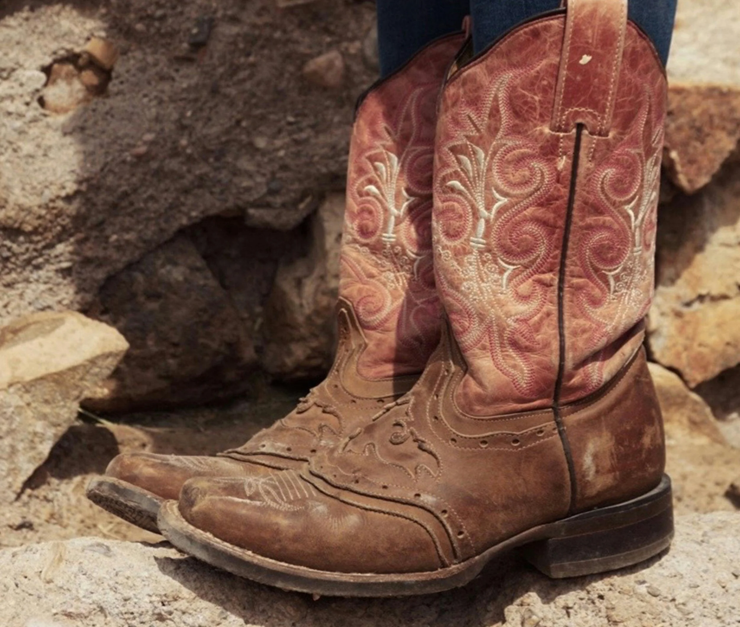A pair of old, worn cowboy boots with intricate embroidery, placed on rocky ground against a stone wall.