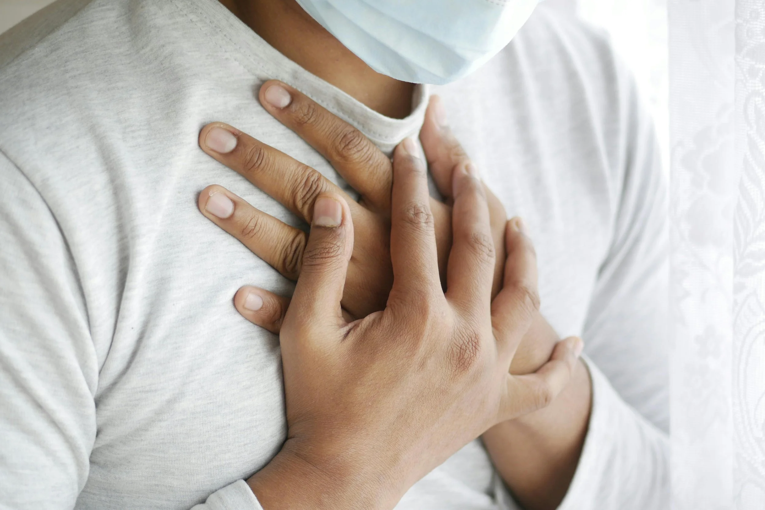 Person wearing a face mask with hands placed on their chest, suggesting they are experiencing chest pain or discomfort.