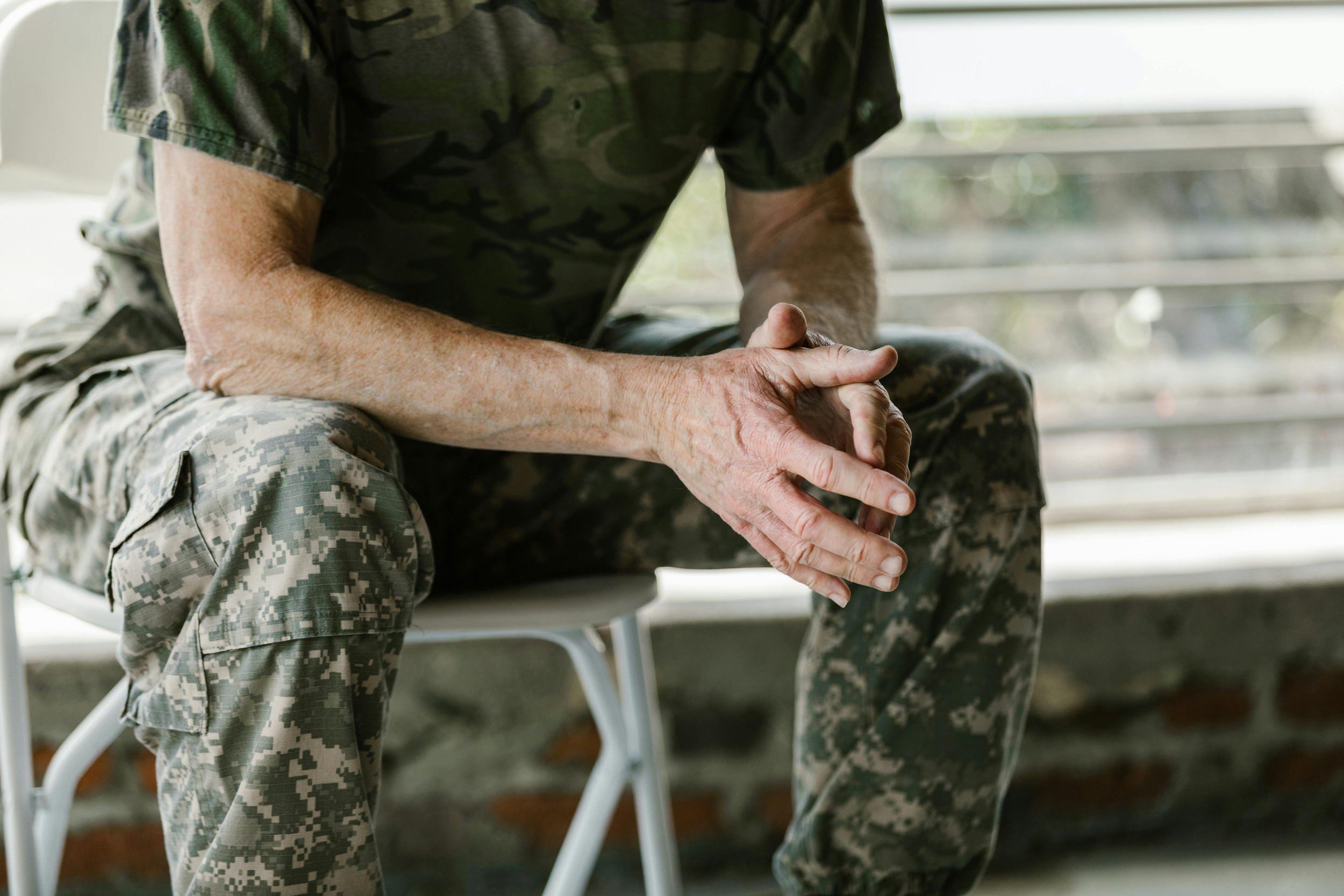 A person in camouflage military pants and a camo T-shirt sitting on a chair with hands clasped together.
