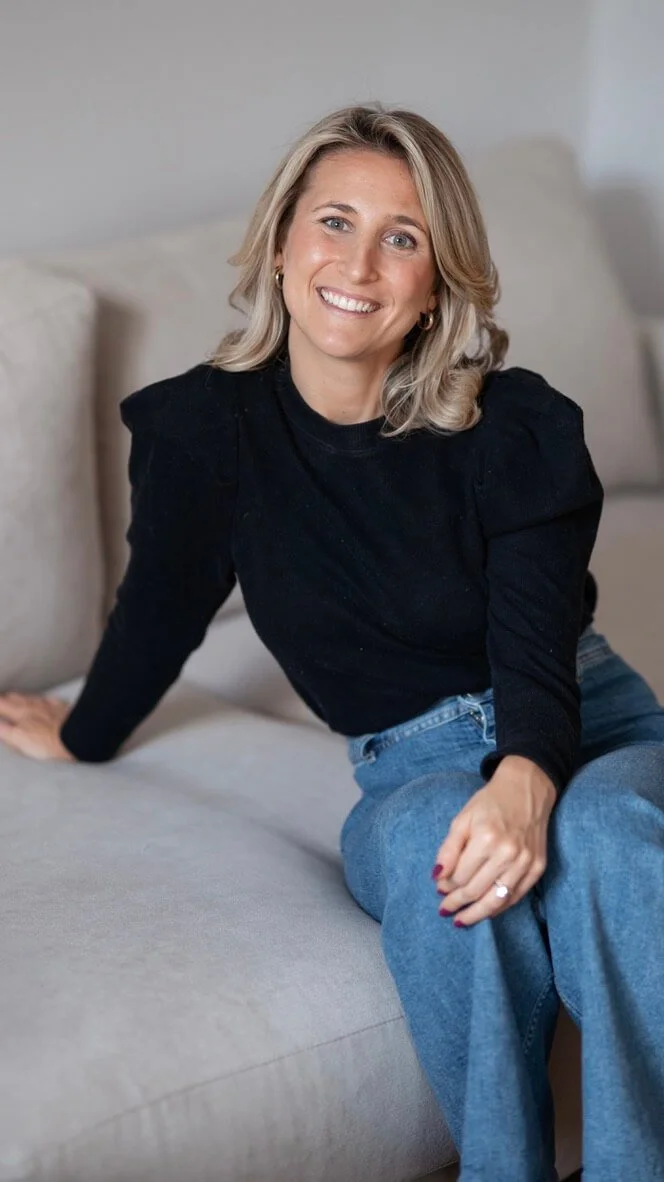 A trauma and anxiety therapist with blonde hair, wearing a black long-sleeve top and blue jeans, sitting on a beige couch and smiling at the camera.
