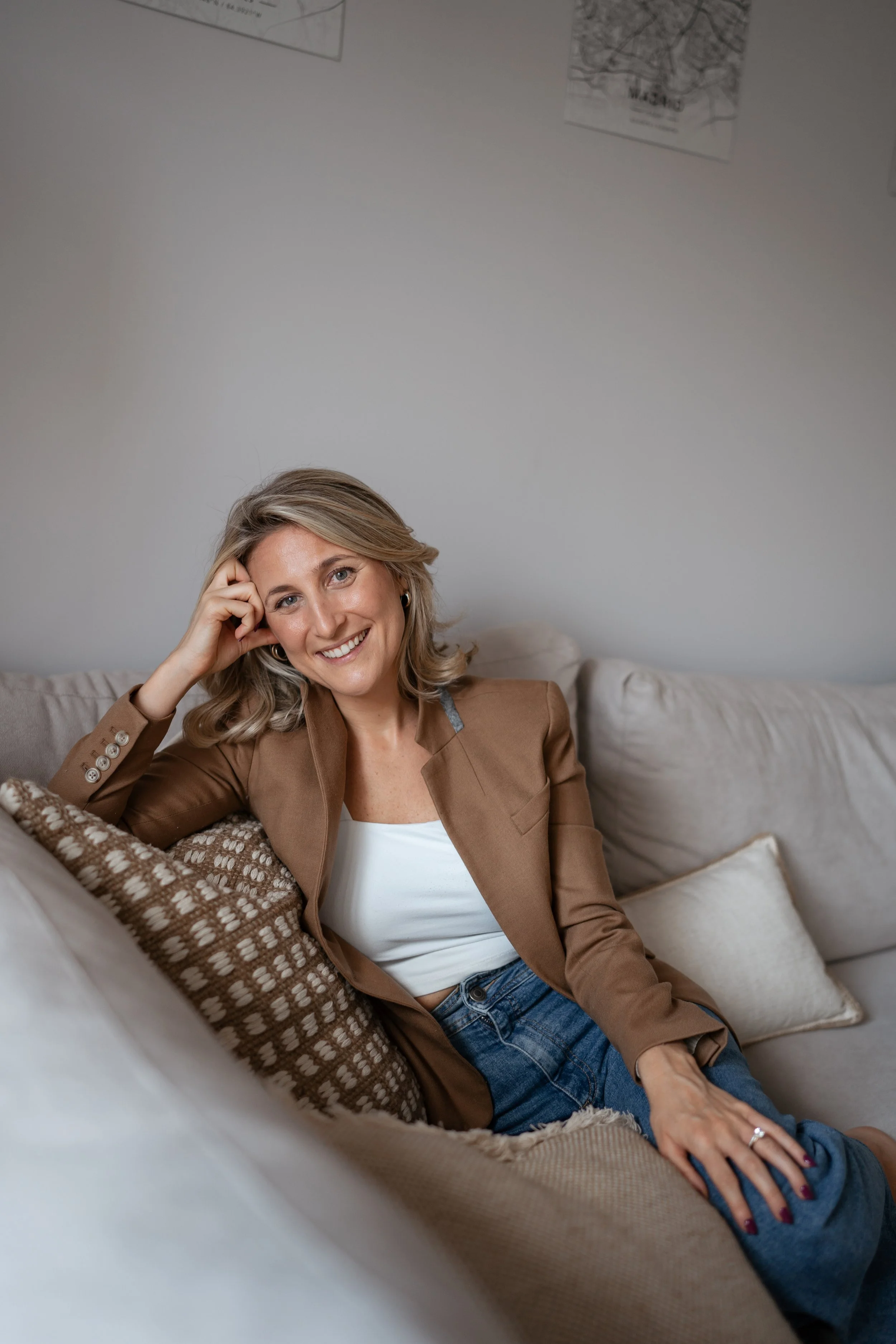 A NYC relationship therapist with blonde hair sitting on a beige sofa, smiling and looking at the camera. She is wearing a brown blazer over a white top, with blue jeans, and is resting her hand on her head.
