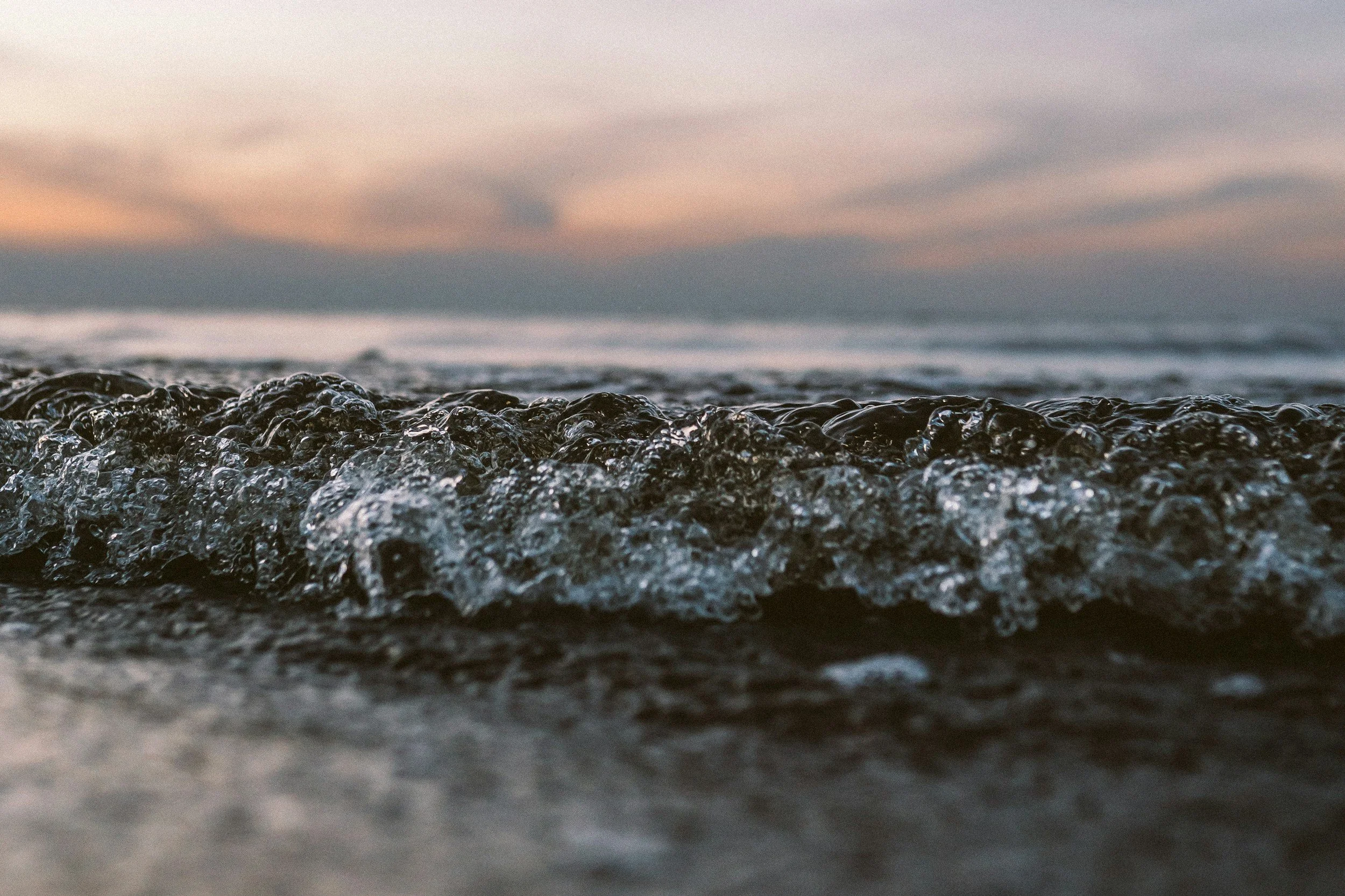 Close-up of ocean waves surfacing on a beach with a sunset in the background.