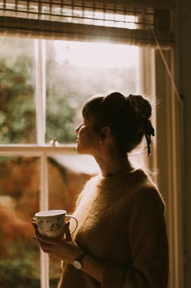 Girl with bun in a sweater holding a coffe cup looking out the window reflecting after anxiety therapy