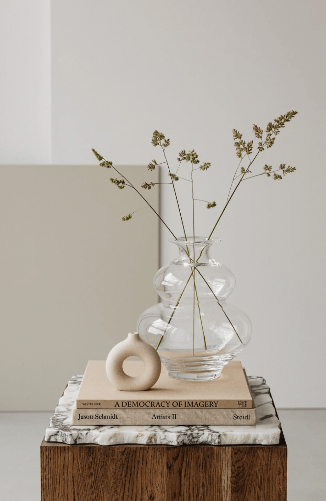 Decorative arrangement with a clear glass vase holding dried green stems on top of two stacked books with a small ceramic vase in front of the books, all placed on a wooden table with a marble top against a light-colored wall.