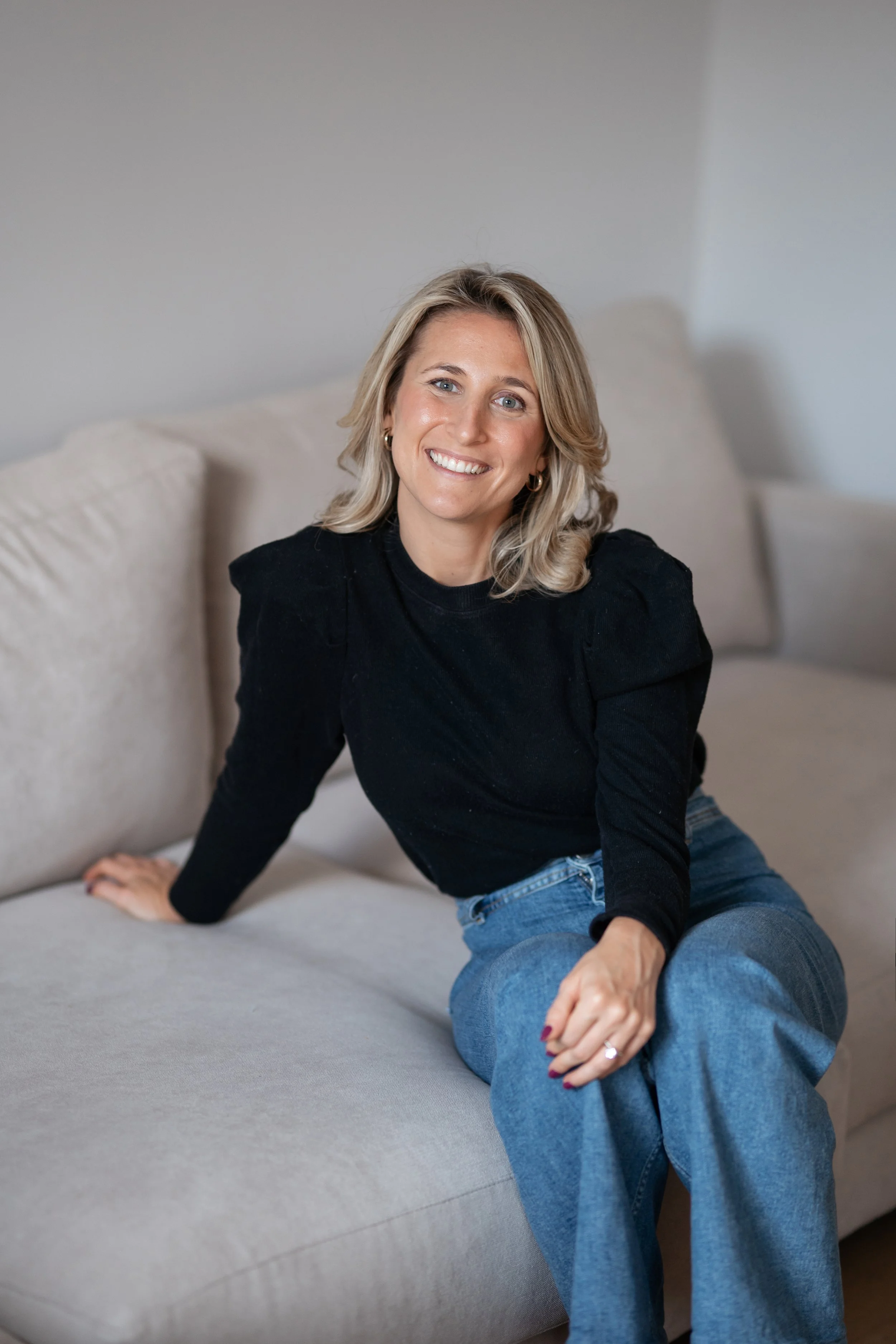 A woman with blonde hair, wearing a black top and blue jeans, sitting on a gray sofa with a beige pillow in the background, smiling at the camera in a well-lit room.