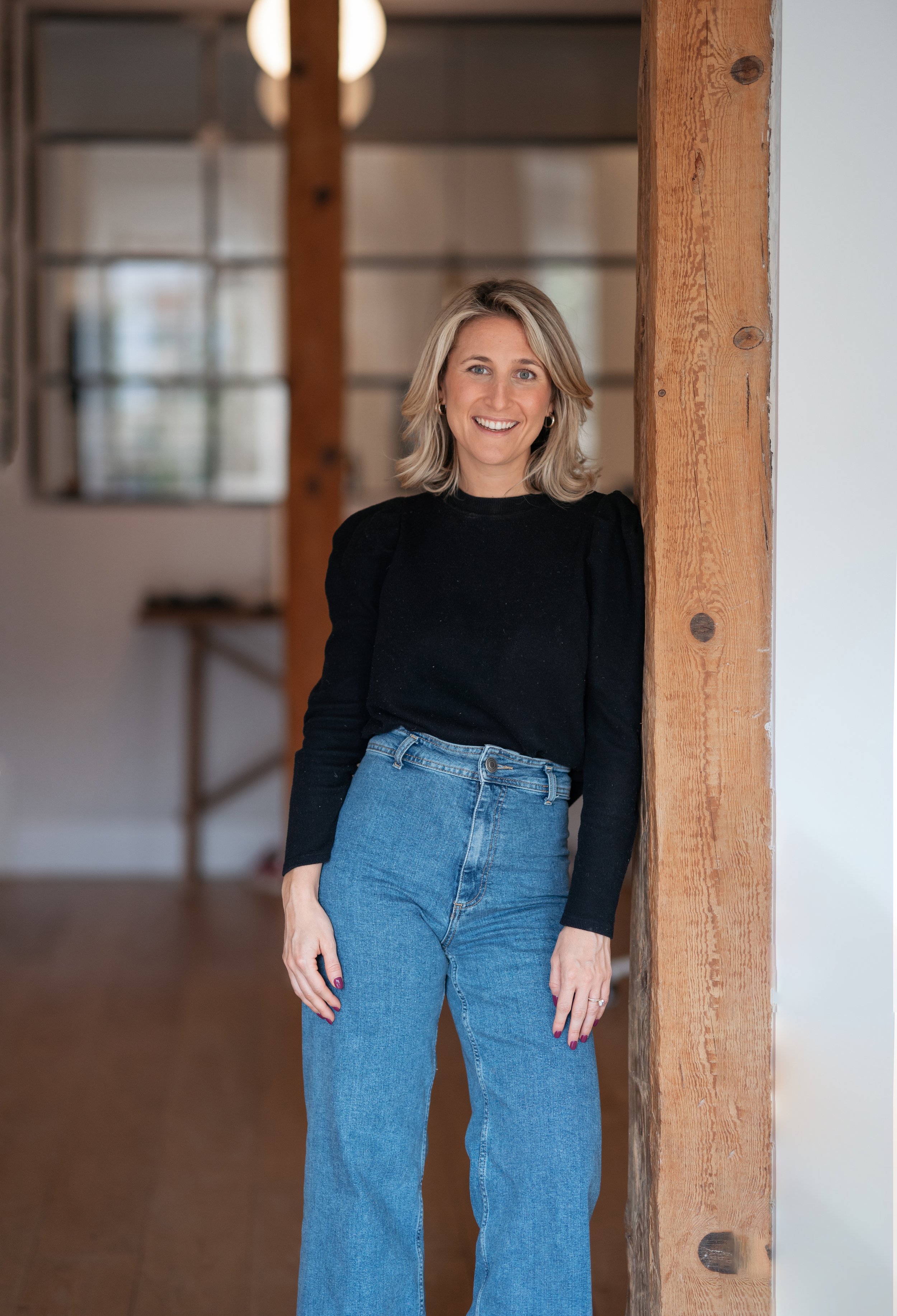 A woman with blonde hair, blue eyes, wearing a black top and blue jeans, smiling and leaning against a wooden beam in a modern interior space.