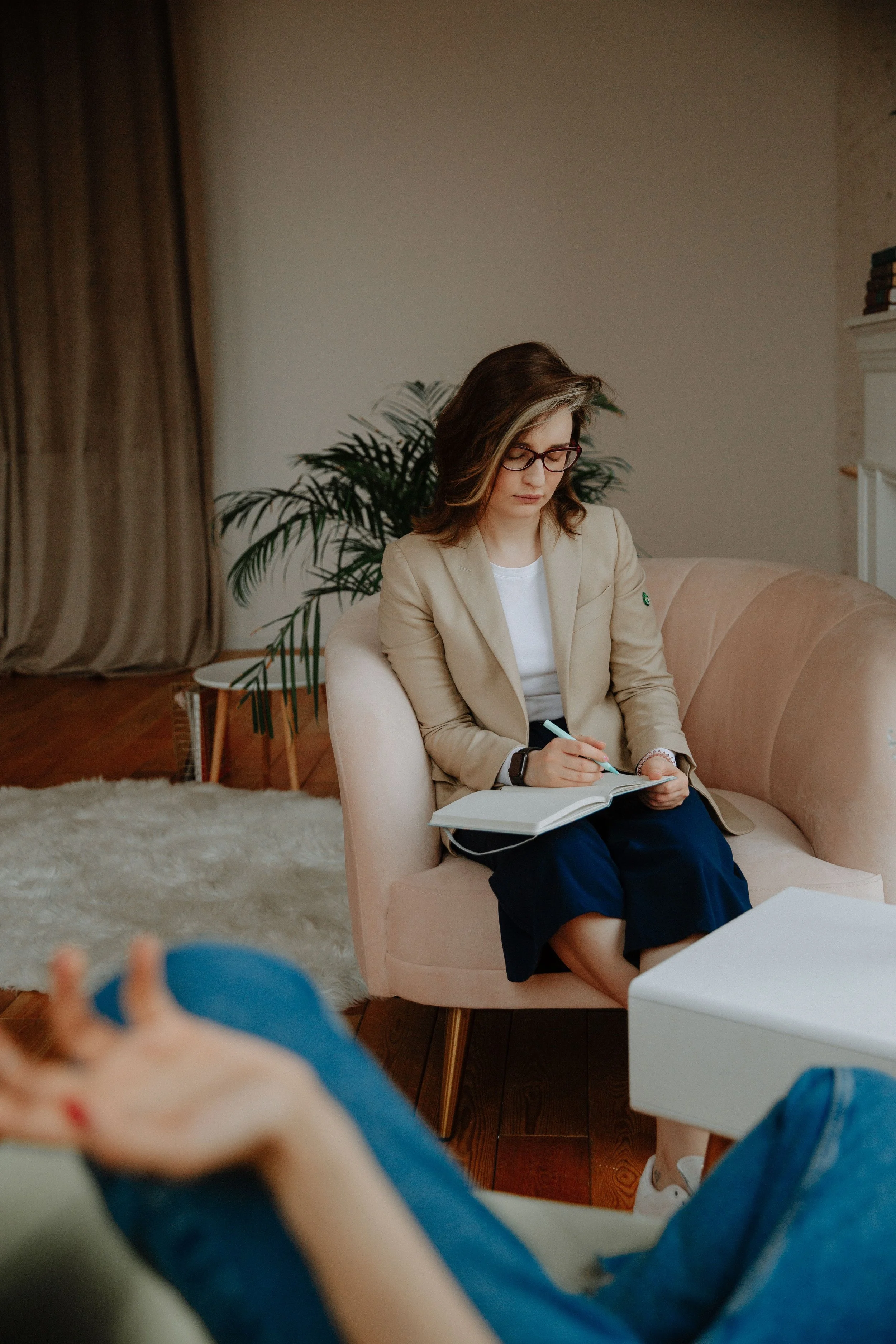 A woman with brown hair, glasses, beige blazer, and white shirt sitting on a beige sofa writing in a notebook.