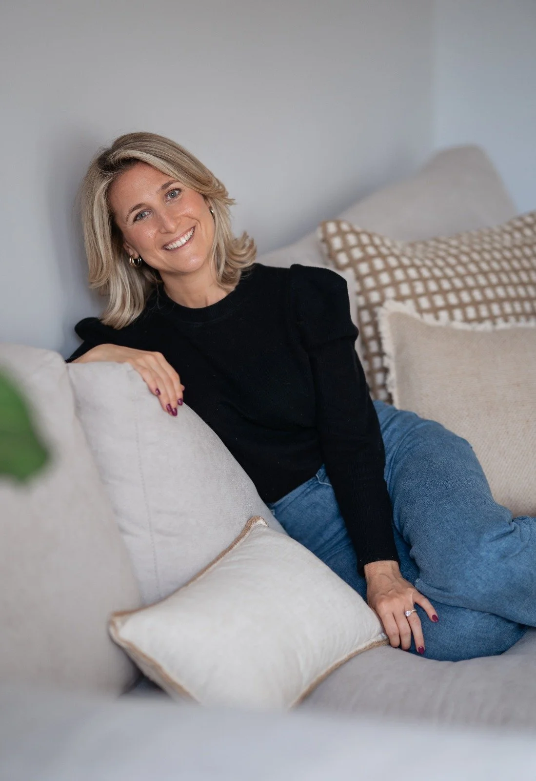 A smiling woman with blonde hair sitting on a light-colored couch surrounded by beige and patterned throw pillows.