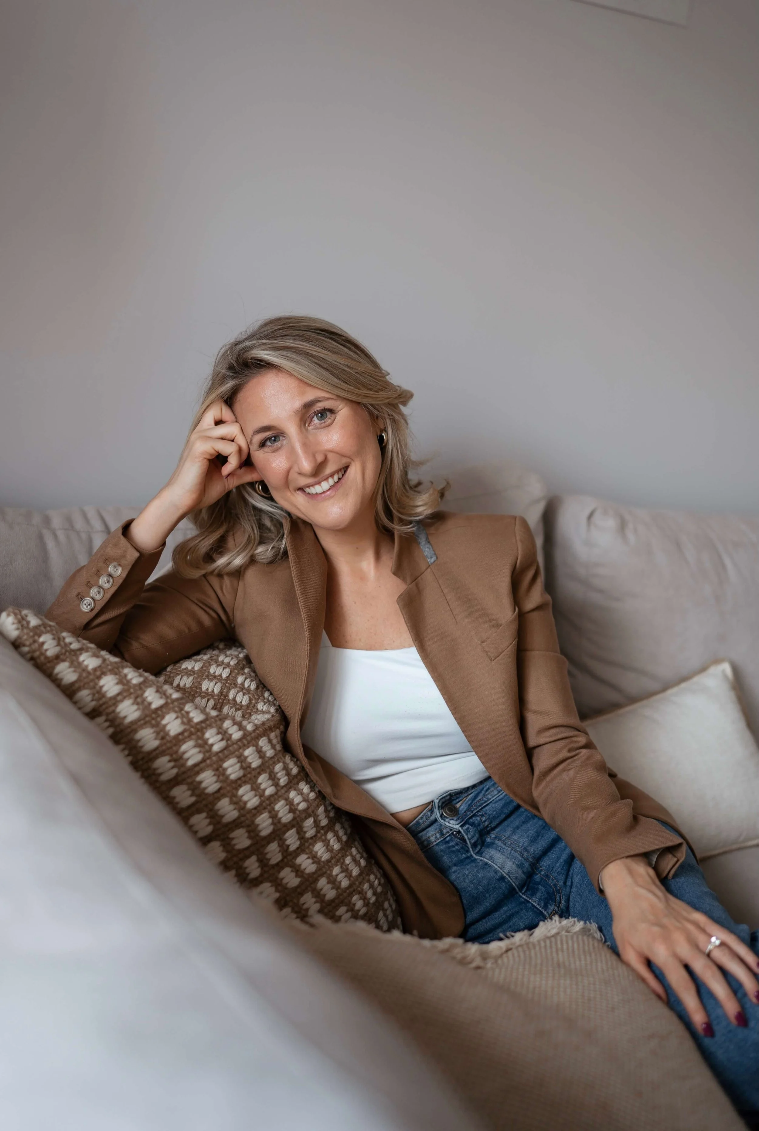 Blonde woman trauma therapist in brown blazer with hand on forehead smiling while sitting on cream couch