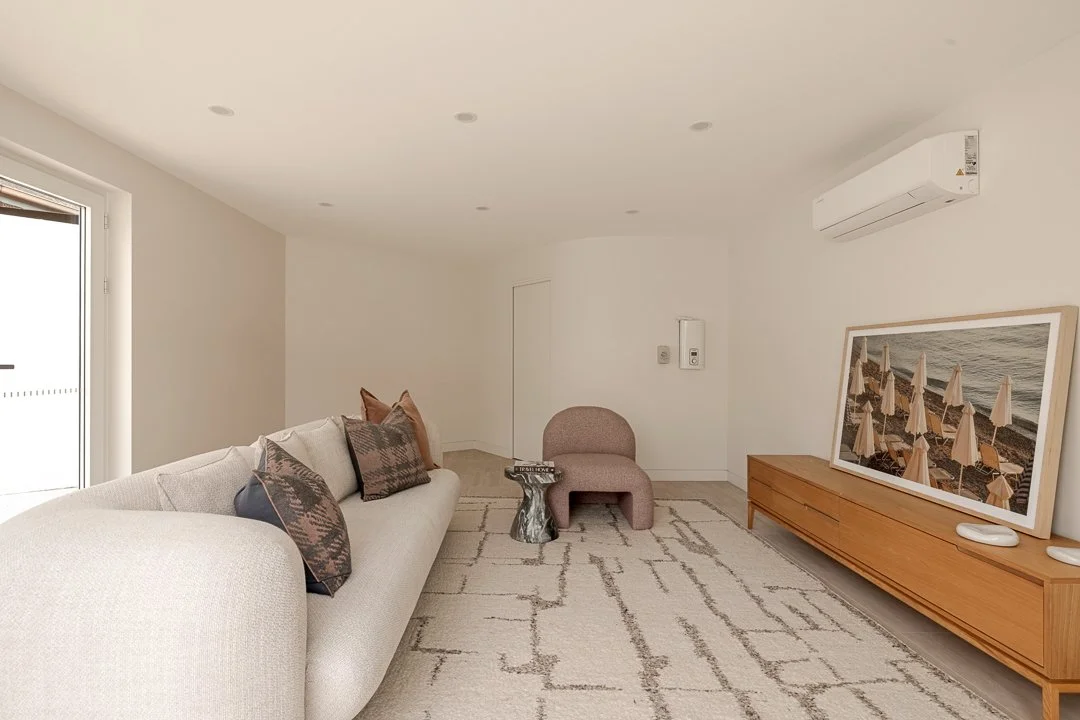 Modern living room with a light-colored couch, decorative pillows, a small pink chair, a wooden console with a framed beach scene photo, a textured rug, and a window with a door, minimal decor, and neutral tones.