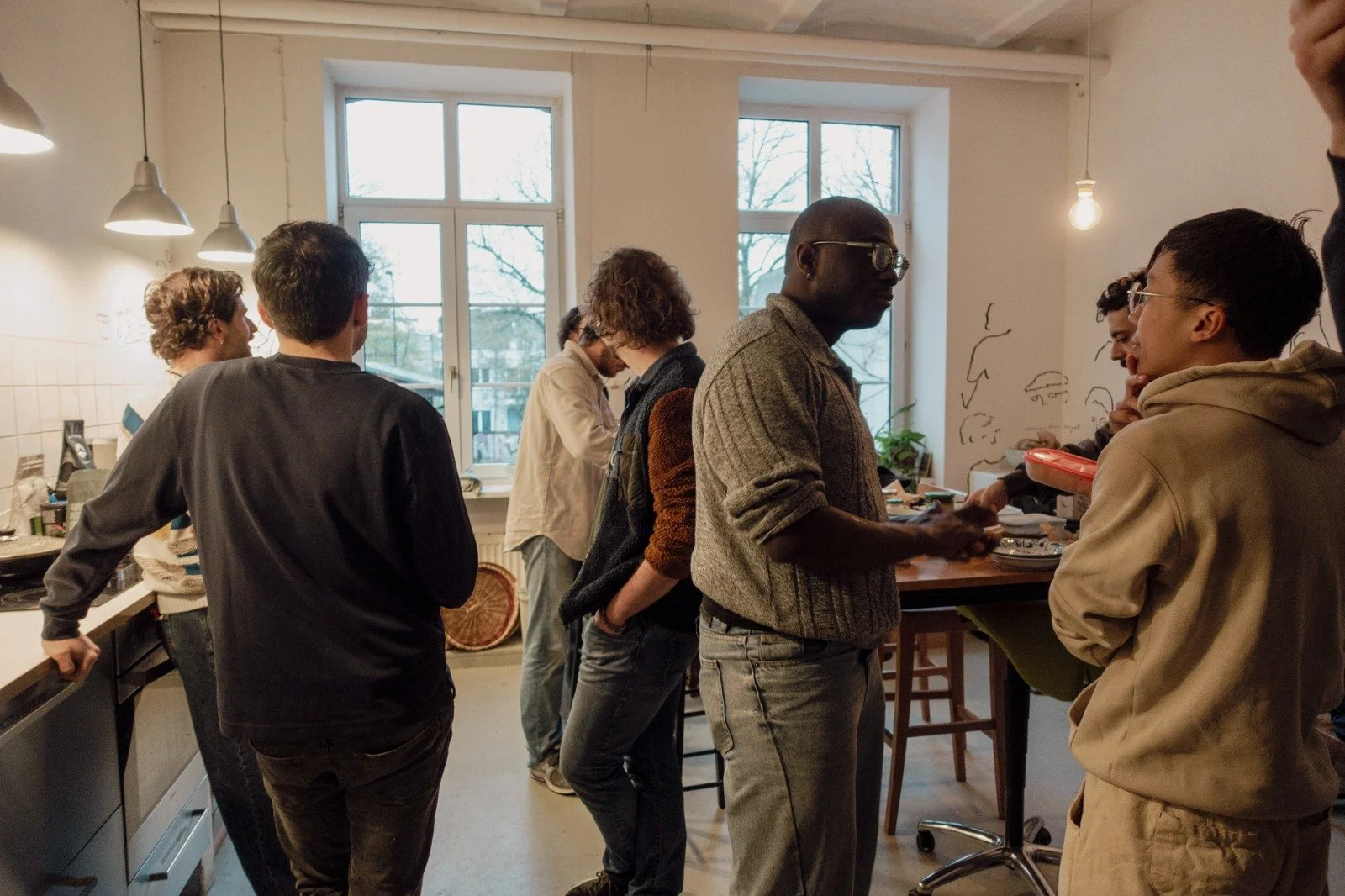 Group of people gathered in a kitchen, standing and talking near a counter with a window in the background.
