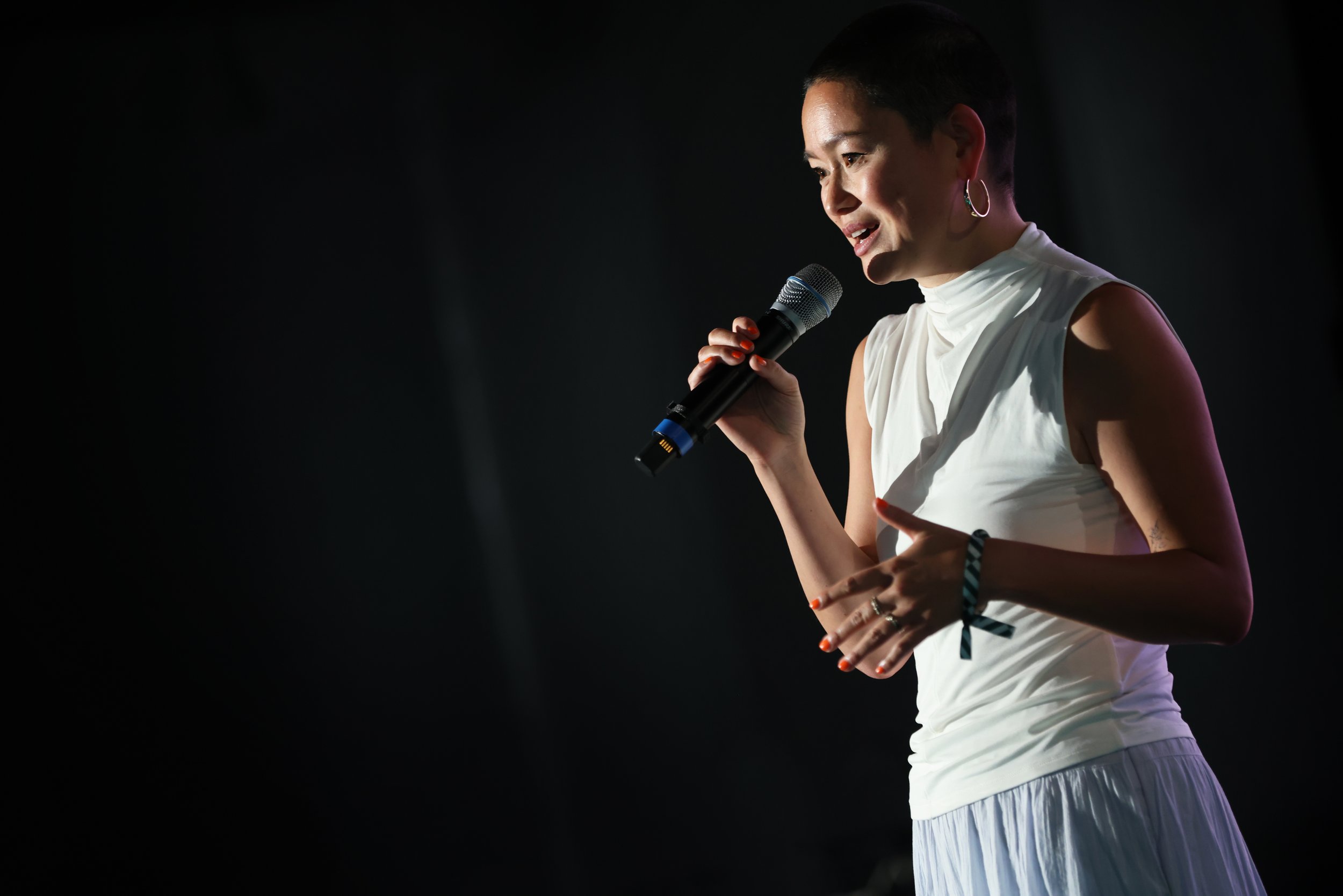 Woman with short dark hair wearing hoop earrings and a sleeveless white top holding a microphone on stage.