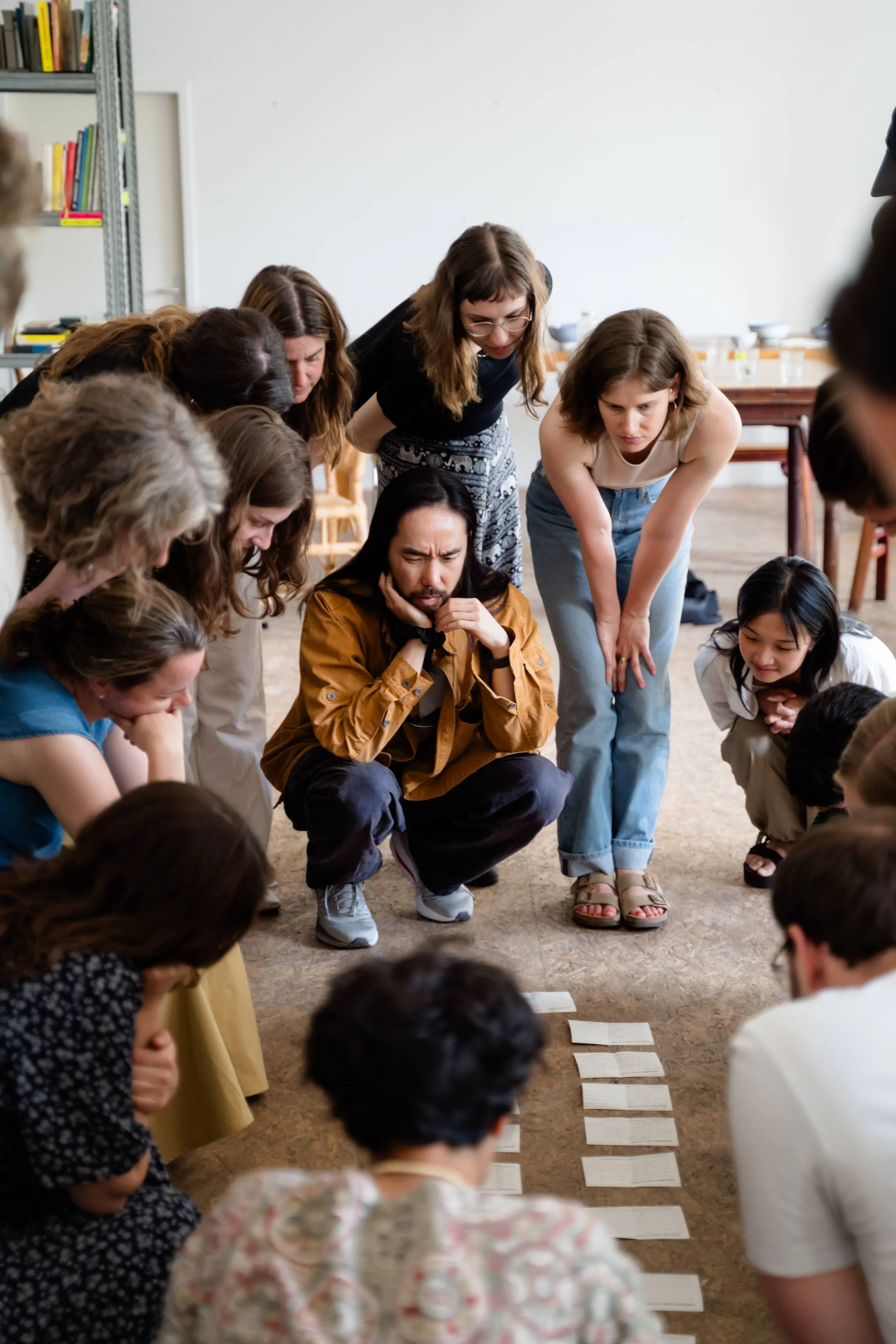 Group of people, mostly women and one man, gathered closely, looking down at a sequence of papers or cards laid out on the floor, in a room with bookshelves and tables.