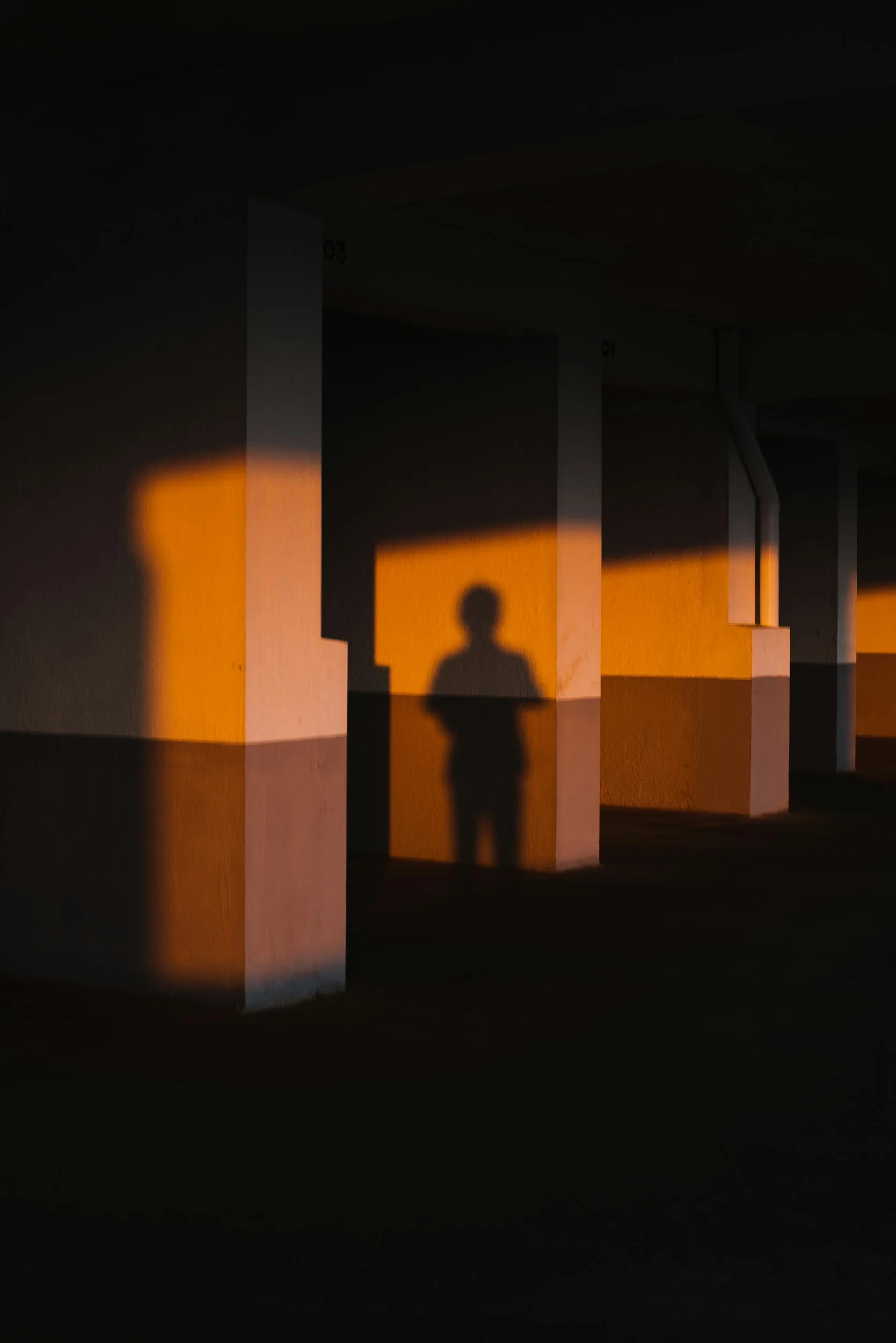 Shadow of a person on a white column, illuminated by warm sunlight, in an empty parking garage.