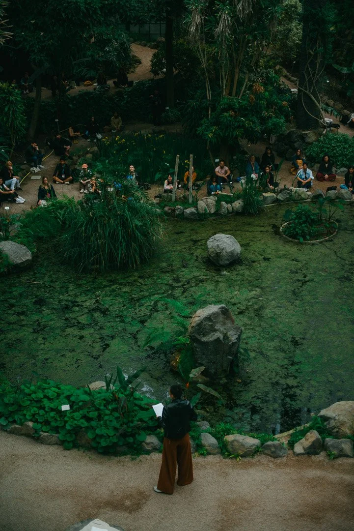 People sitting outdoors around a pond with rocks and lush greenery, some reading or looking at papers, in a park or garden setting.
