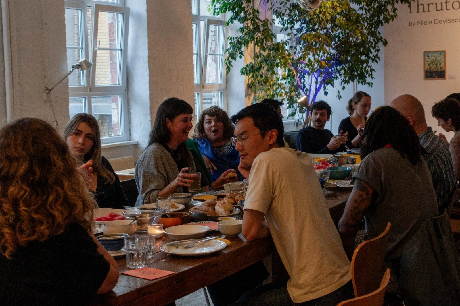 A group of people sitting around a long wooden table enjoying a meal and conversation in a well-lit room with large windows and a potted plant.